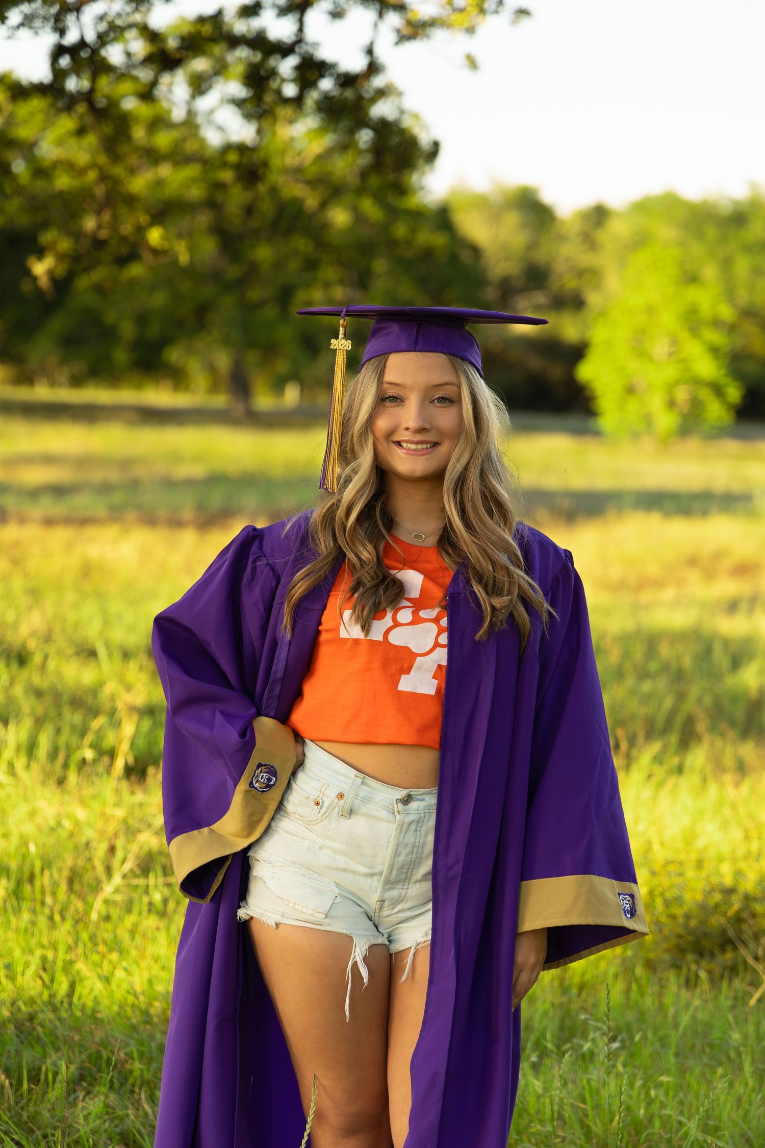 Smiling graduate in purple cap and gown with orange Clemson shirt and denim shorts posing in sunny green field.