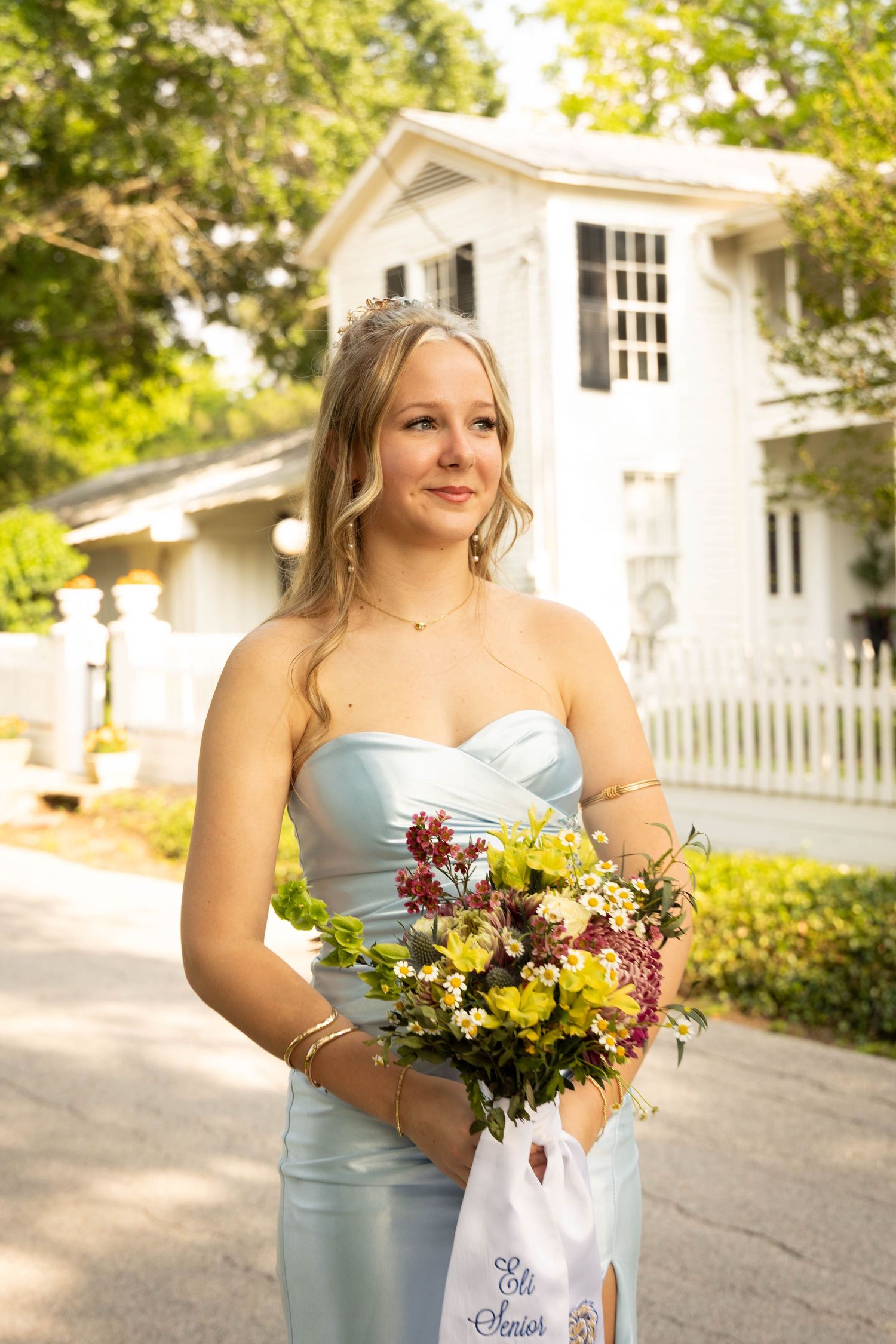 Blonde woman in light blue strapless gown holding wildflower bouquet standing outdoors near white house.