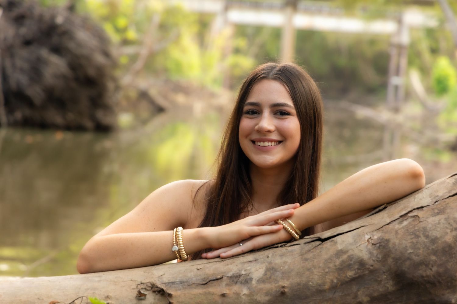 Young woman with long brown hair smiling outdoors near a river, wearing gold bracelets, leaning on a log.