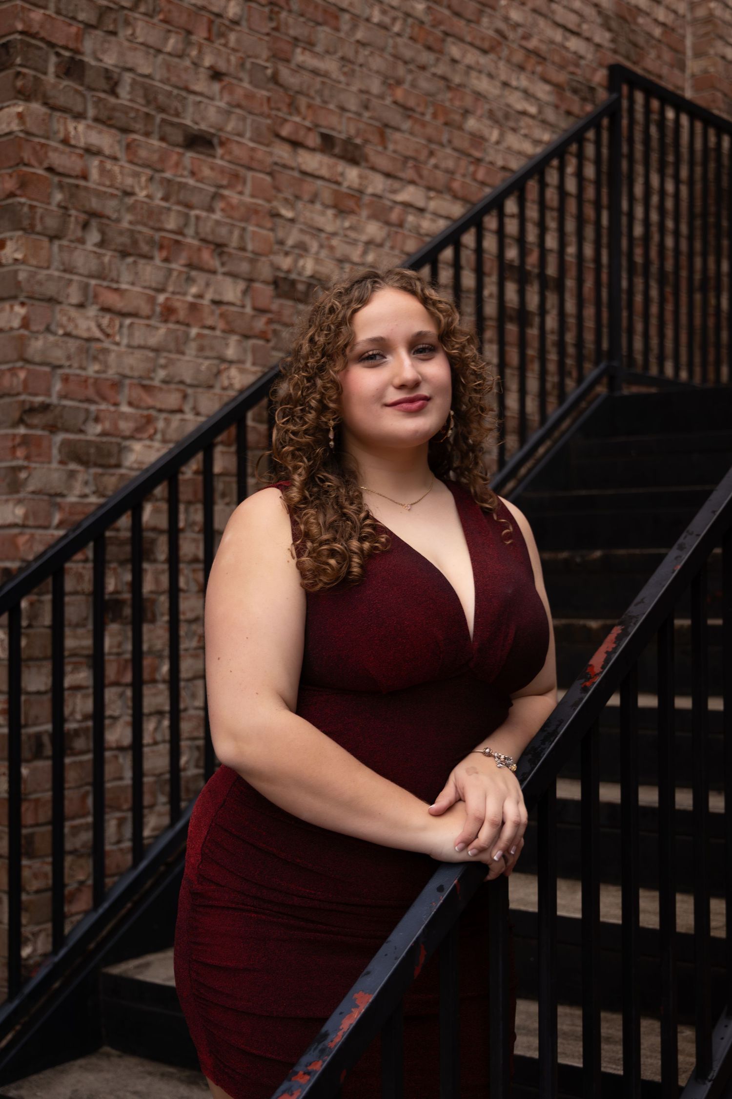 Woman in burgundy dress posing on black metal staircase against rustic brick wall.
