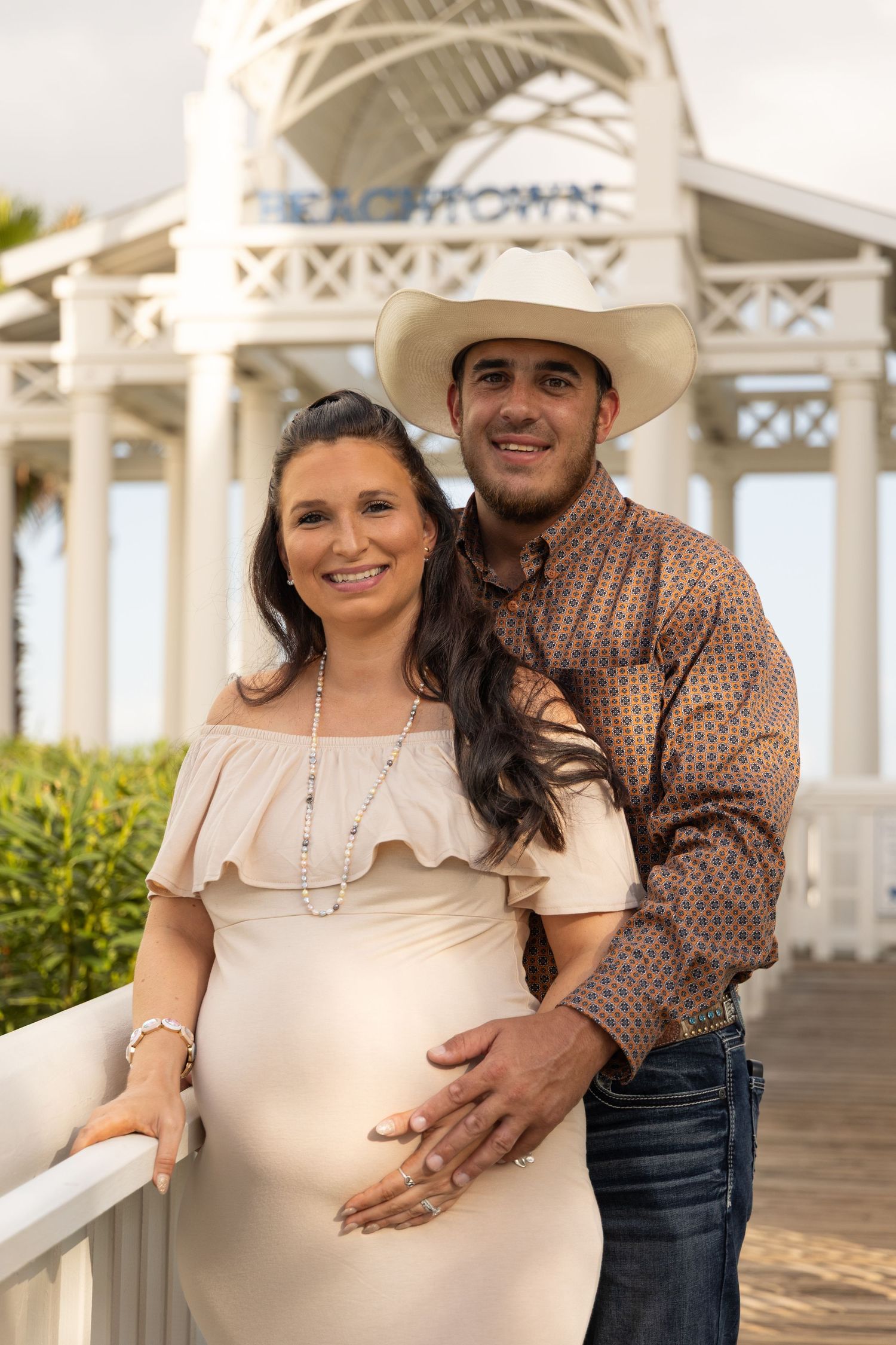 Happy pregnant couple posing at Beachtown gazebo, woman in white maternity dress, man in cowboy hat and western shirt.