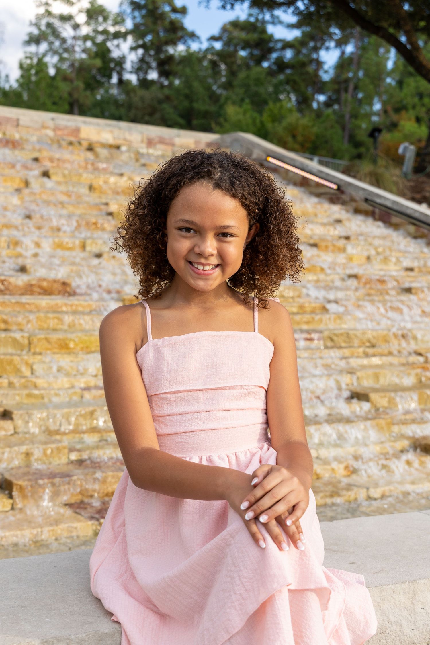 Young girl in pink dress with curly hair smiles while sitting on stone steps outdoors.