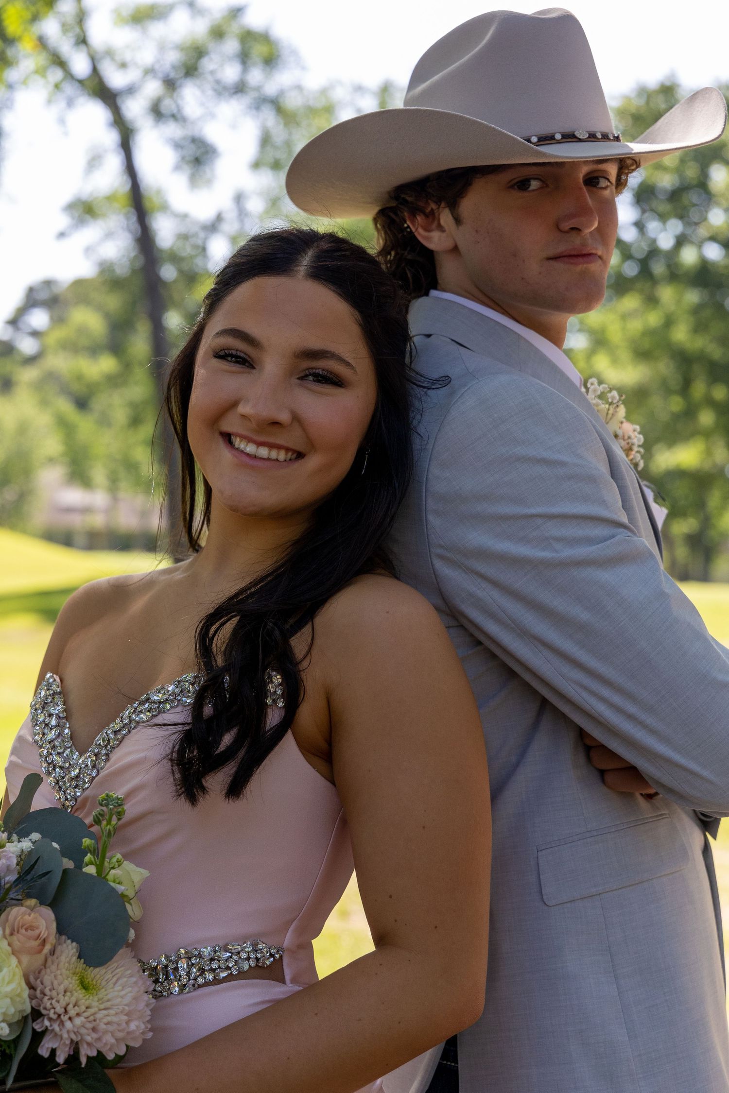 A smiling teen girl in pink prom dress holds flowers beside a young man in gray suit and cowboy hat outdoors.