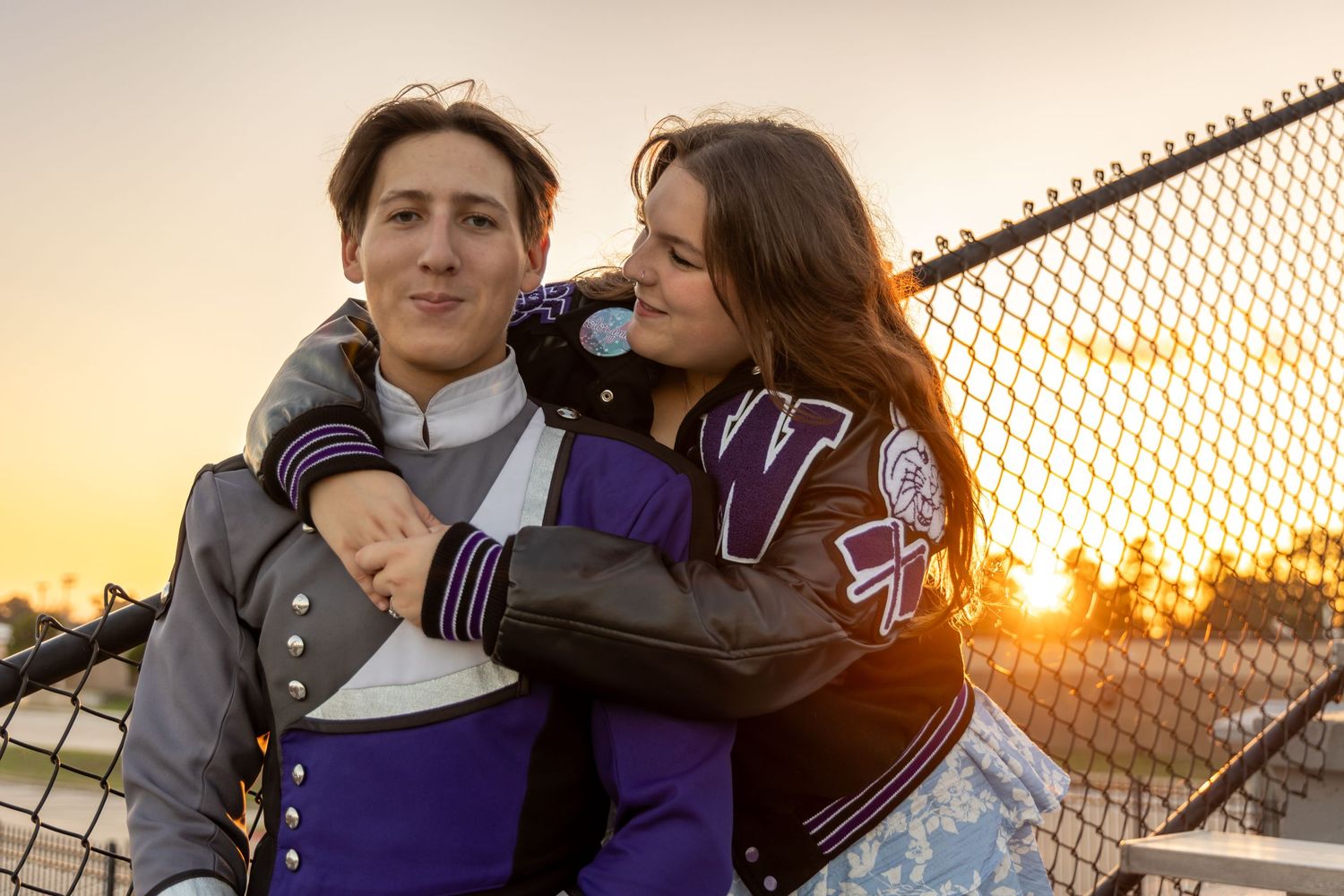 Two people in varsity jackets embrace while standing by a chain link fence during golden hour sunset.