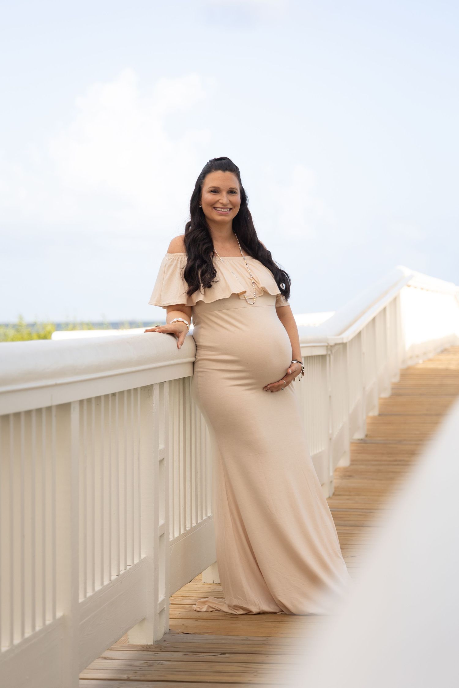 Pregnant woman in beige off-shoulder maternity gown posing on a wooden bridge railing outdoors.