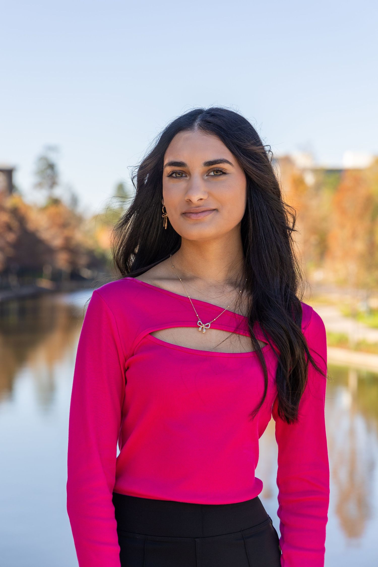Young woman in bright pink long-sleeve top with cutout detail, standing outdoors near a reflective lake with autumn trees.