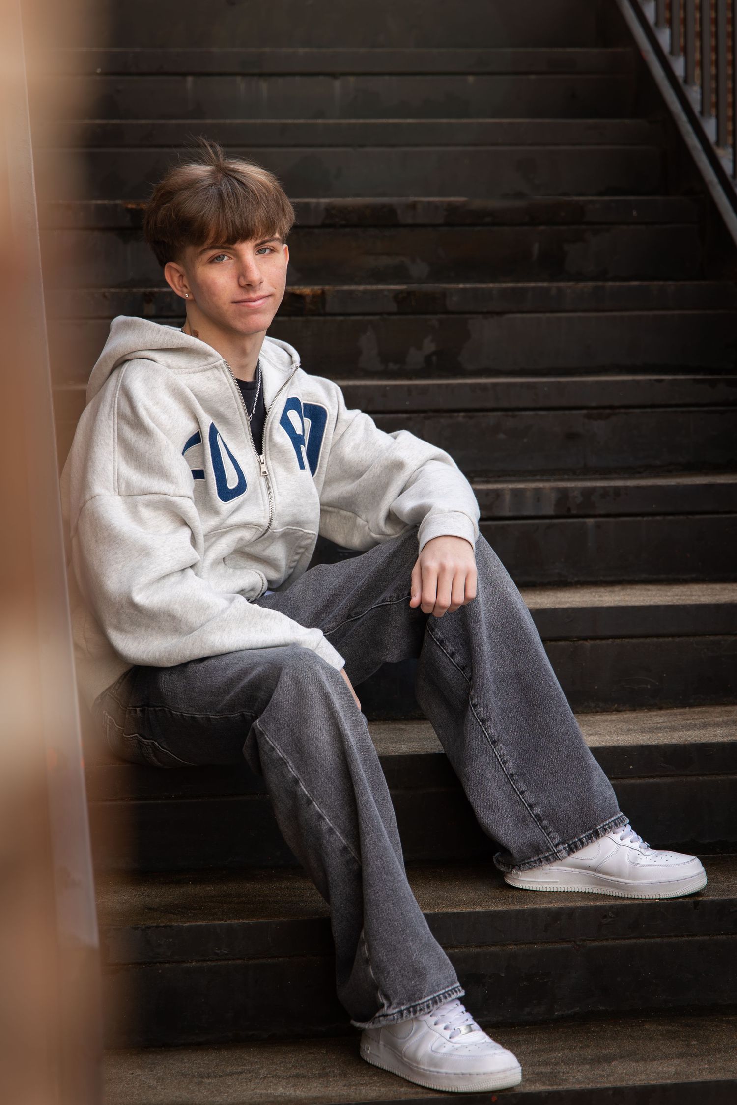 Teen sitting on dark stairs wearing gray hoodie, wide-leg jeans, and white sneakers, posing casually.