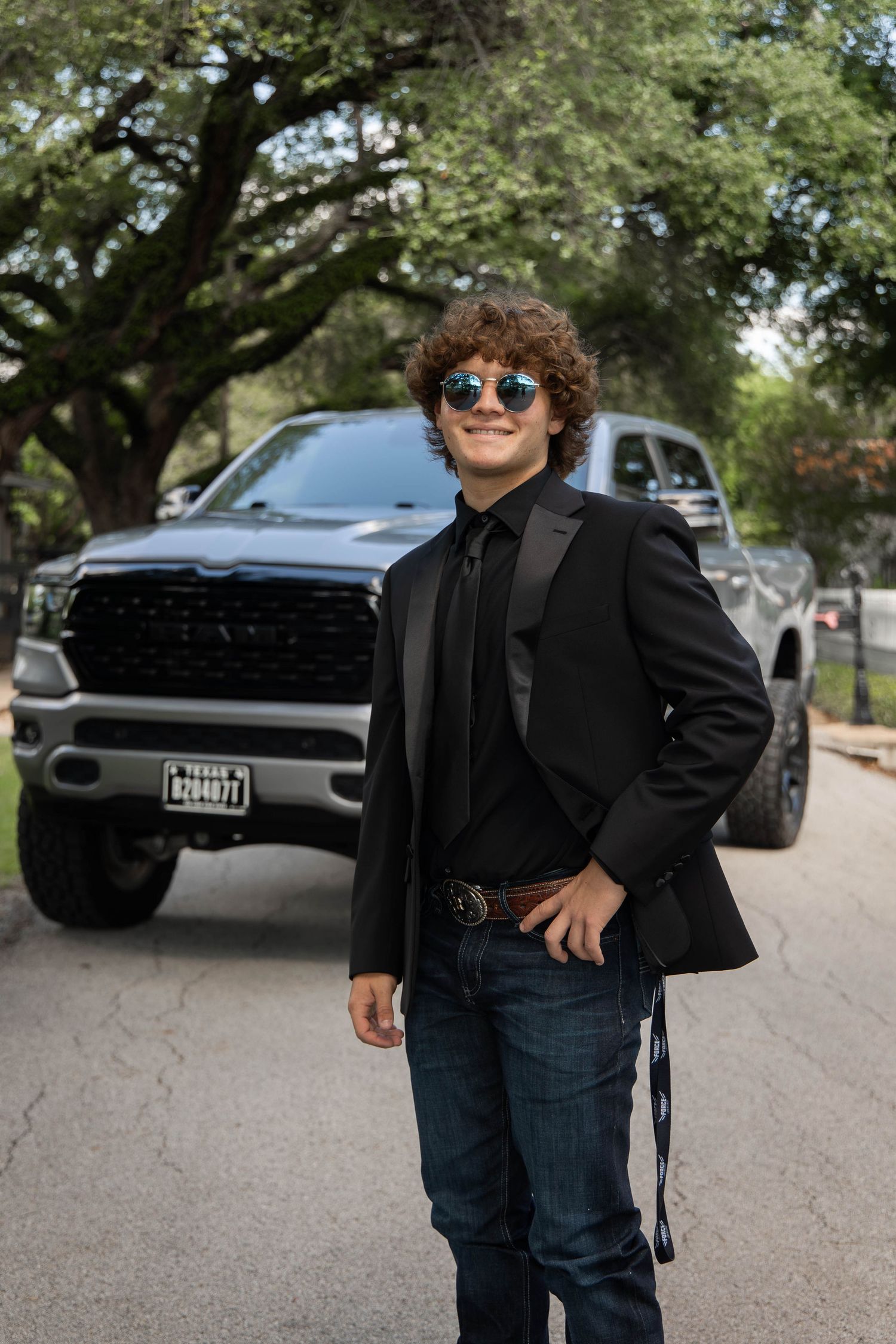 Young man in black tuxedo jacket and sunglasses poses in front of a Ram truck on a Texas driveway.