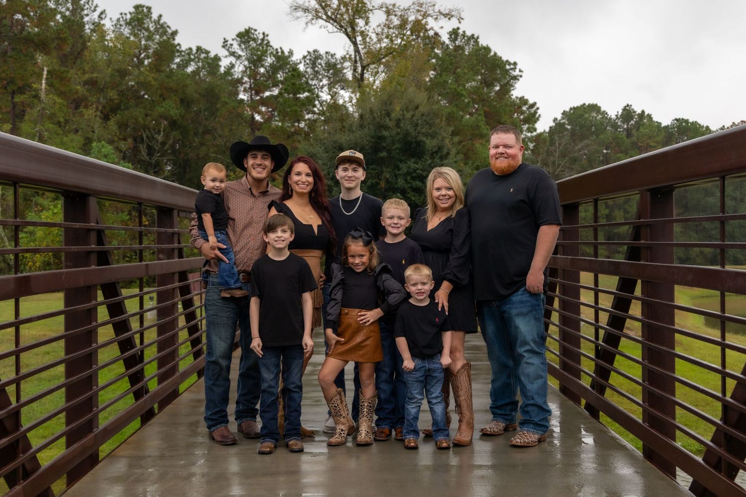 Large family group portrait on a wooden bridge outdoors, dressed in matching black outfits, smiling together.