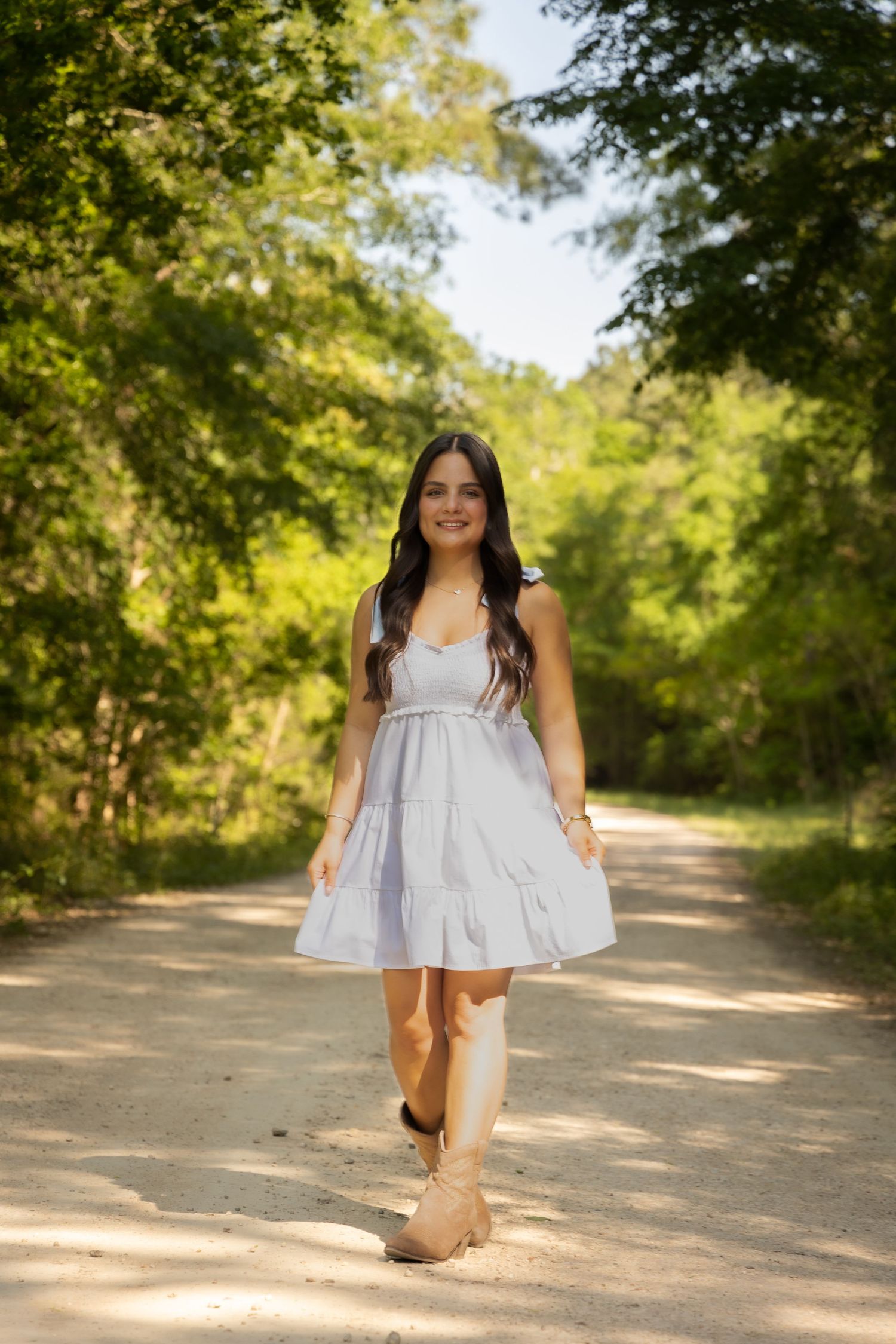 Young woman in white sundress and boots walking on a sunny dirt path surrounded by lush green trees.