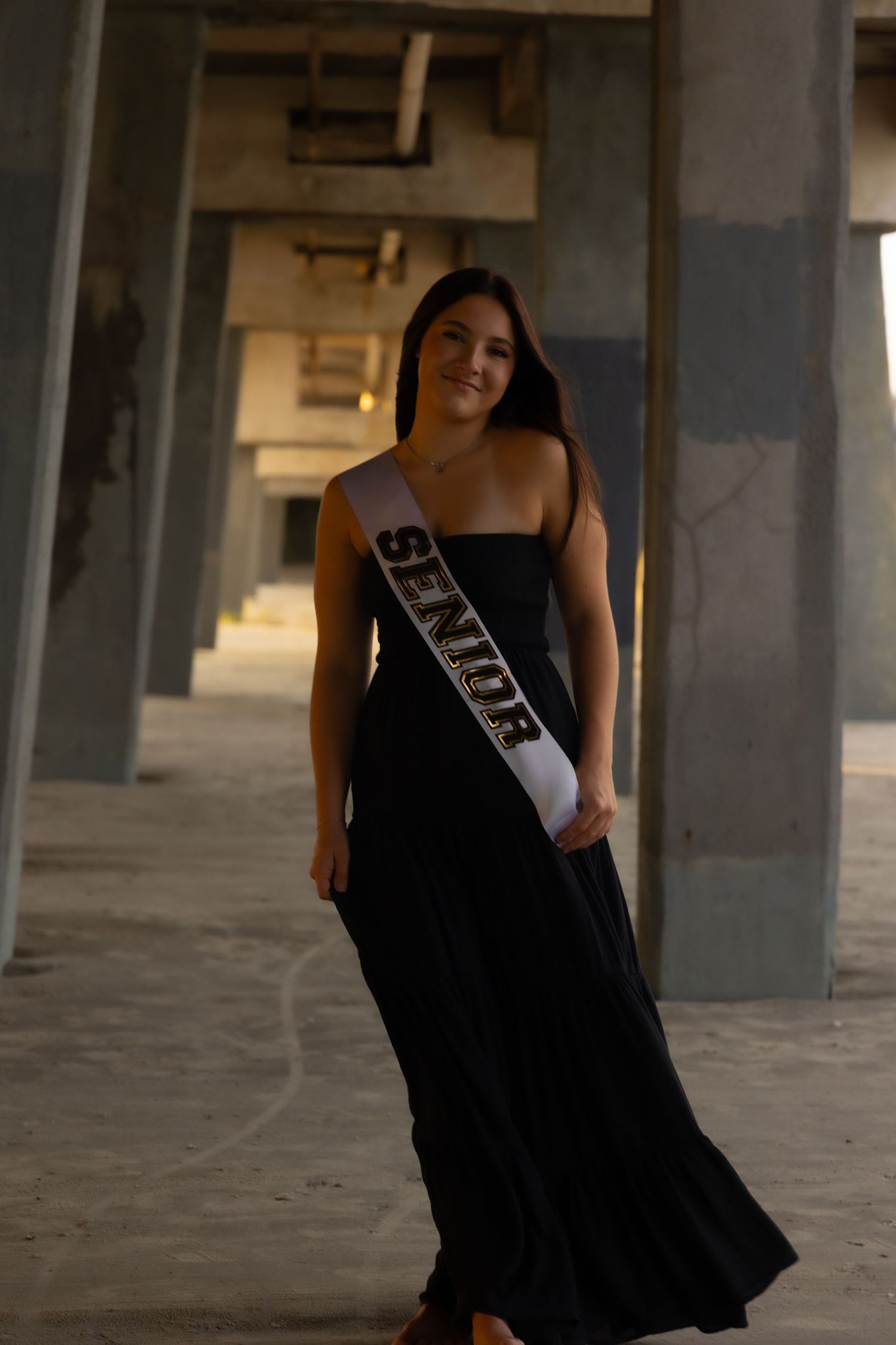 Young woman in black gown wearing a Senior sash poses in an industrial building for senior portraits.