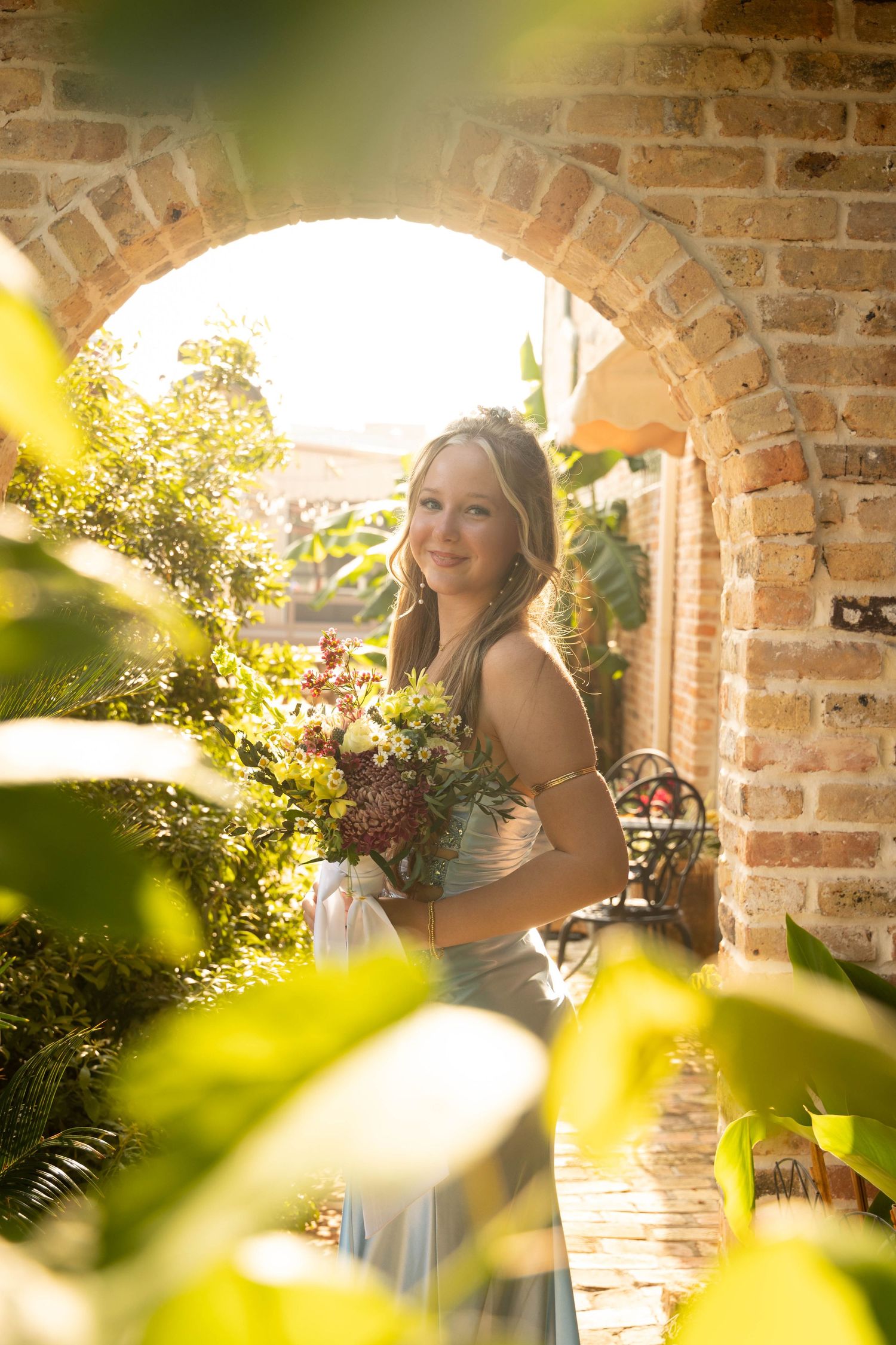 Smiling young woman in blue gown holding bouquet framed by sunlit brick archway surrounded by greenery.