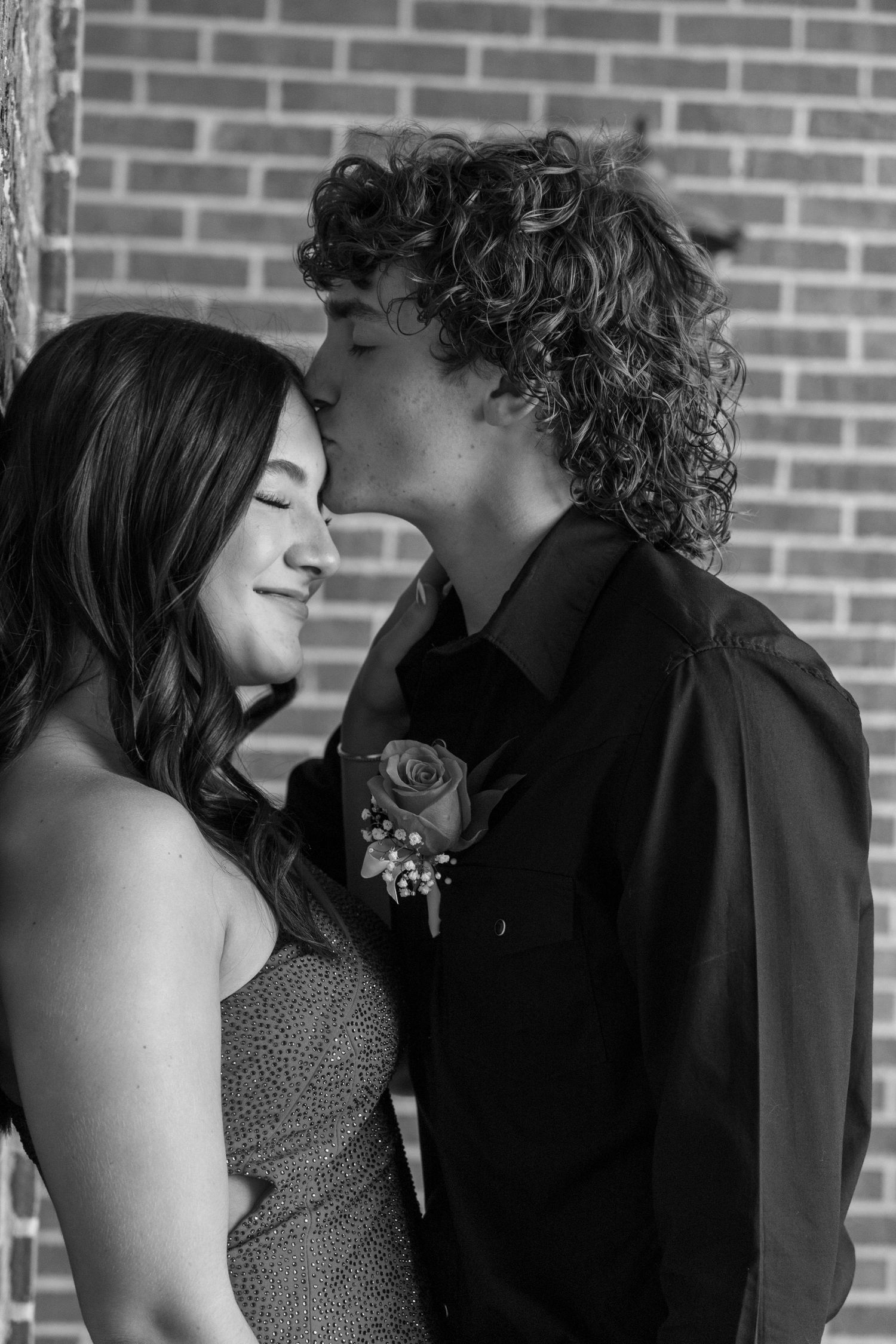 Black and white photo of a young couple at prom, boy with curly hair kissing girl on cheek, rose boutonniere.