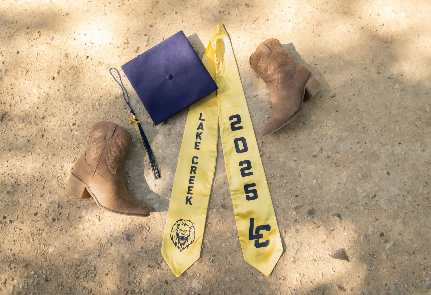 Graduation cap and yellow university sash with class of 2023 text lay in the sand alongside rocks.