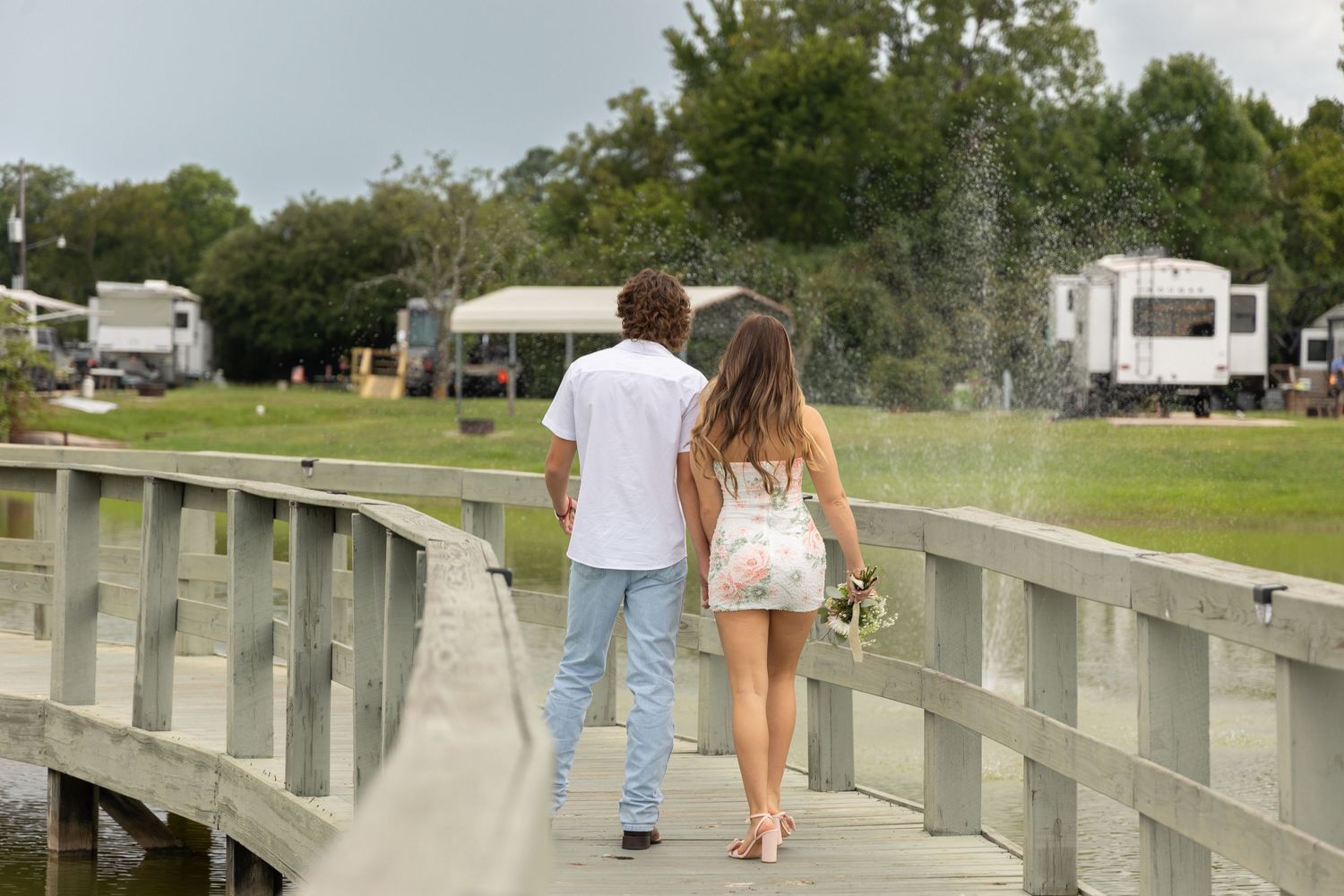 A couple walks hand-in-hand across a wooden bridge surrounded by lush greenery at a campground.