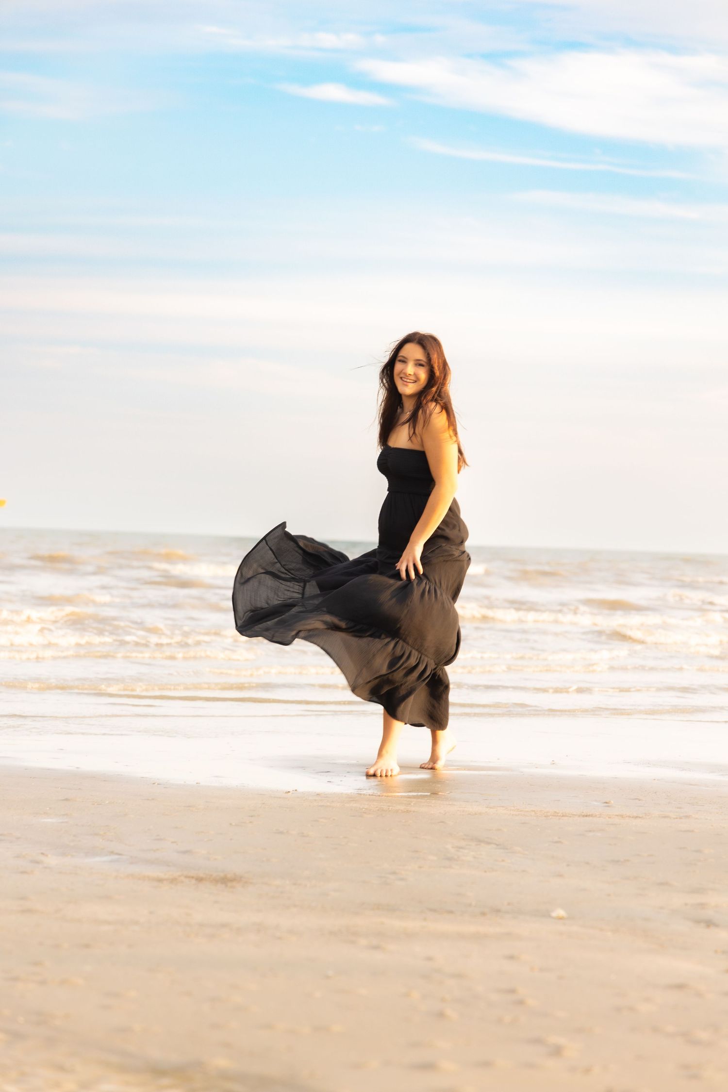 Woman in flowing black dress standing on sandy beach with ocean waves and blue sky backdrop.