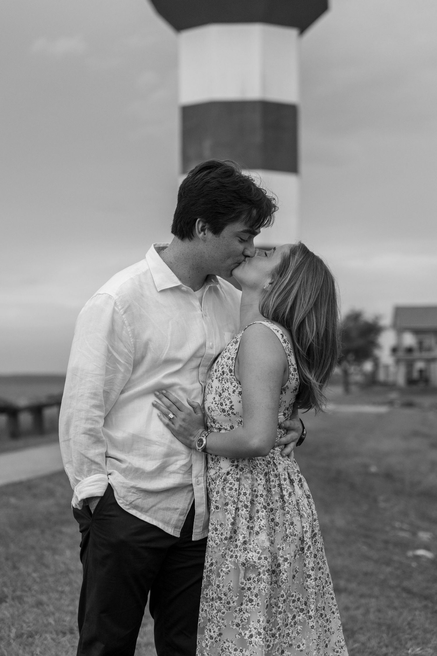 Couple shares a romantic kiss in black and white photo with lighthouse backdrop near the water.