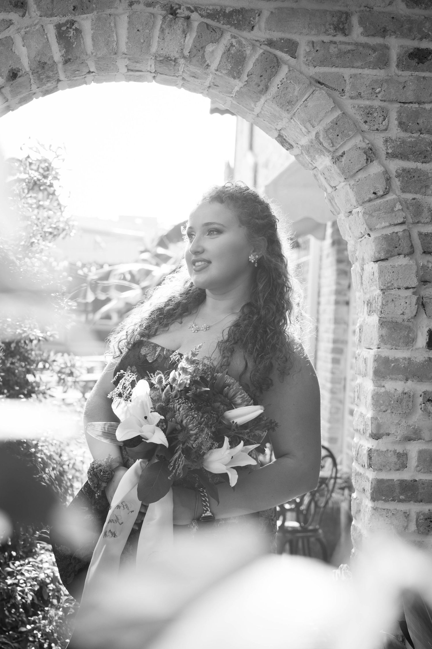 Black and white portrait of curly-haired woman in strapless gown holding lily bouquet under brick arch.