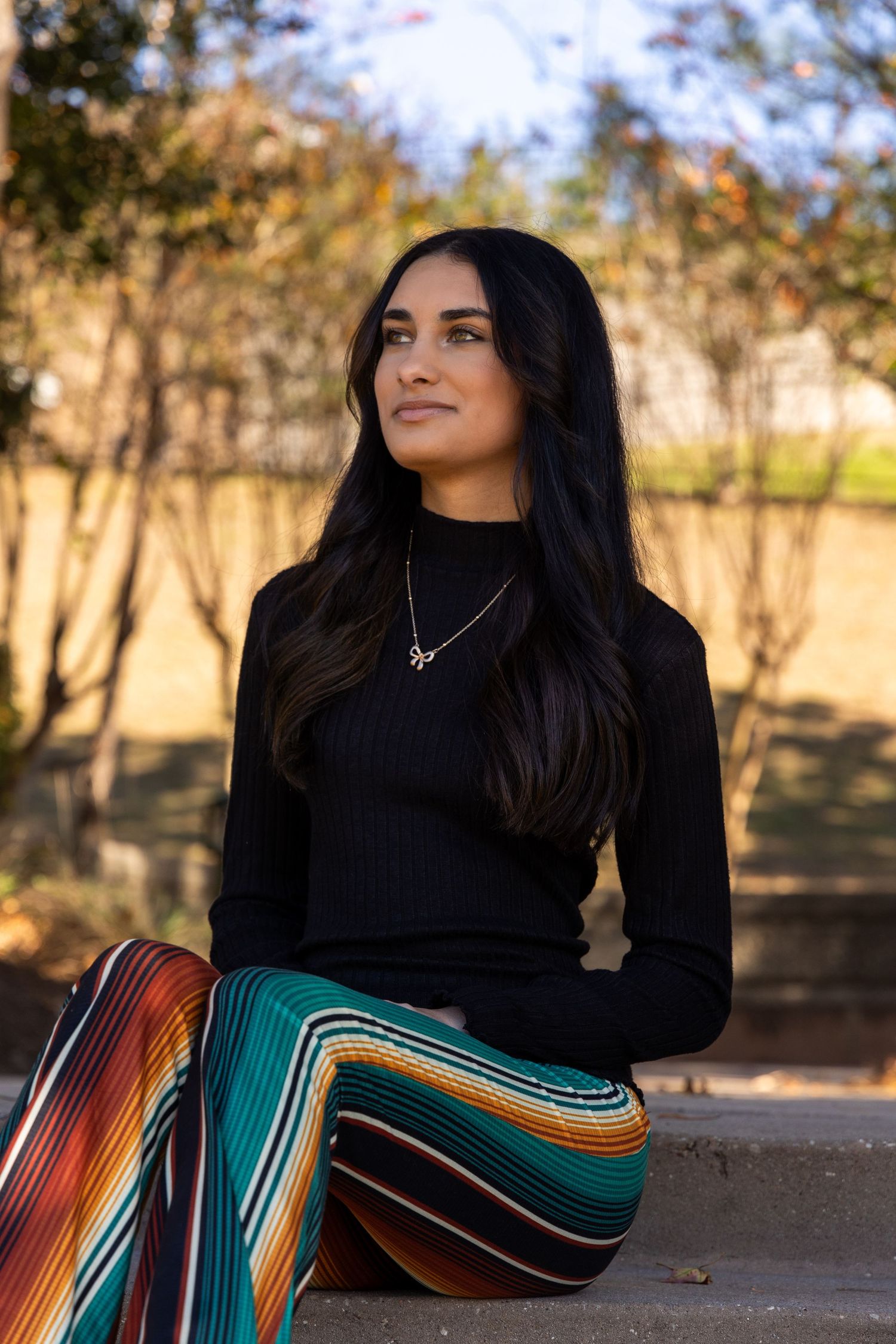 Woman with long dark hair sits outdoors in black turtleneck and colorful striped pants, gazing thoughtfully to the side.