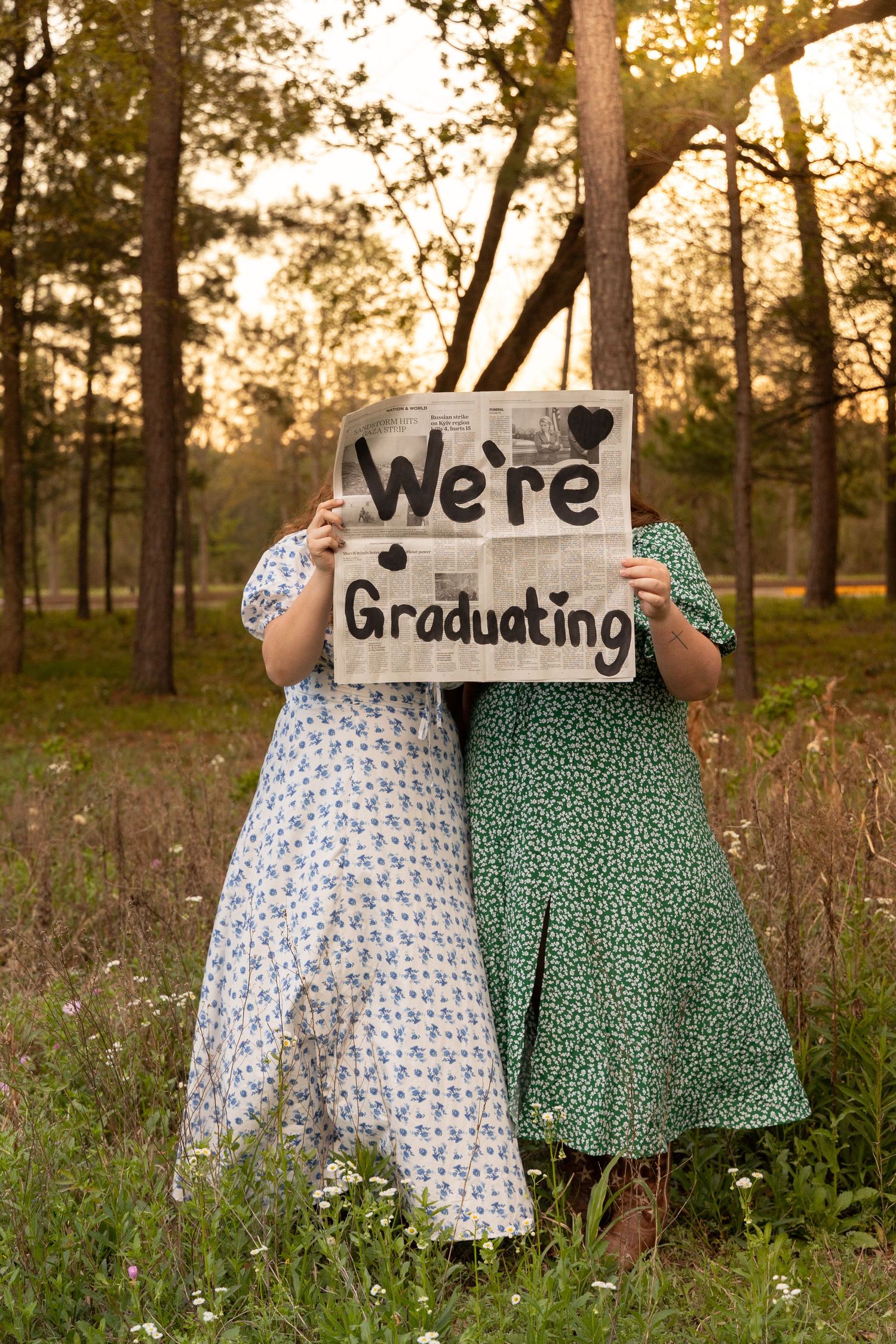 Two graduates in floral dresses hold a handmade cardboard sign reading We're Graduating outdoors at sunset.