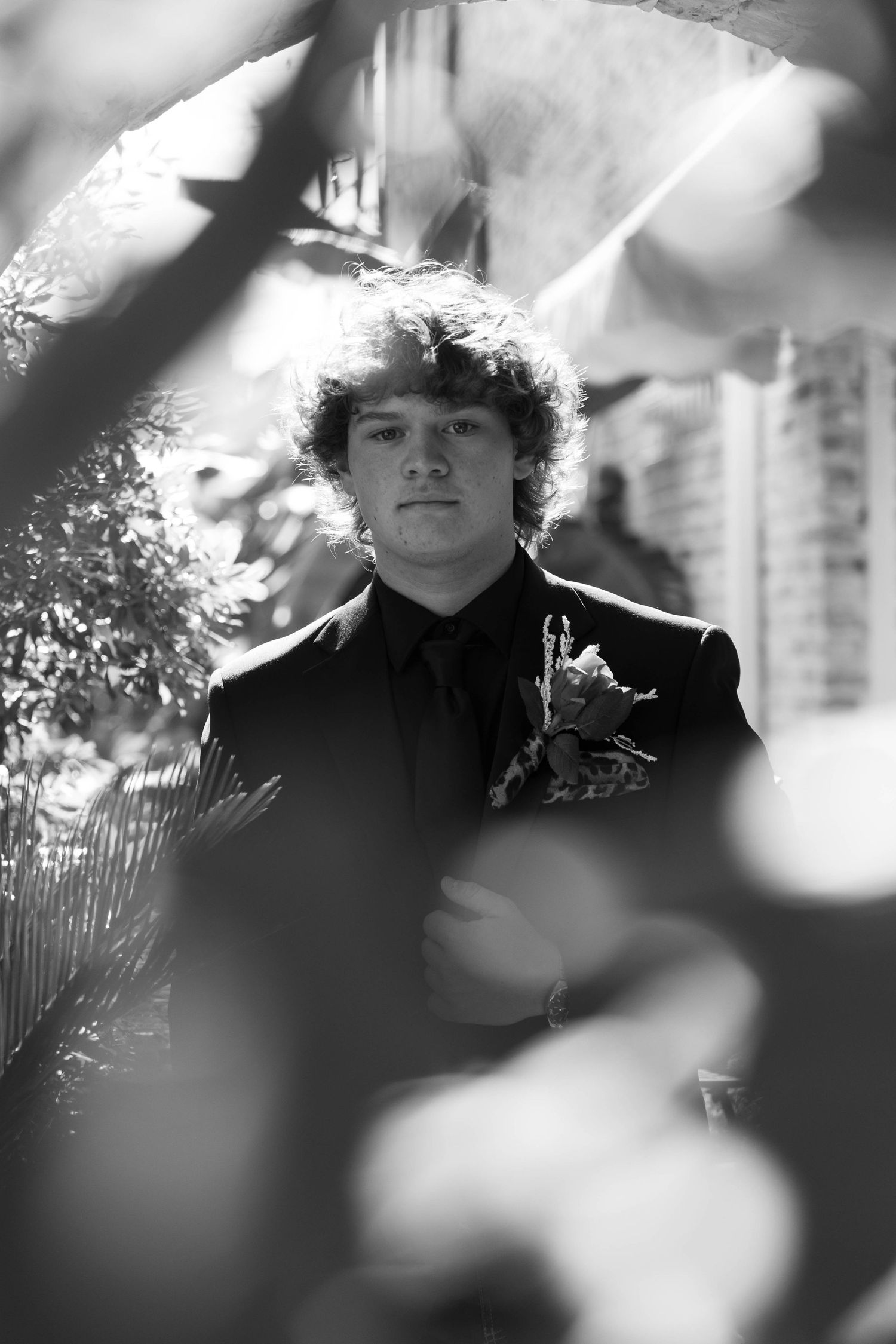 Black and white portrait of a young man in a suit with boutonniere, framed by blurred foliage foreground.