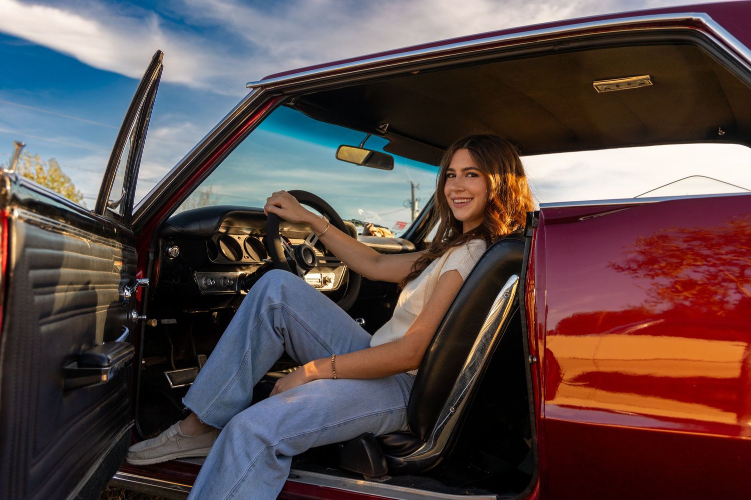 Someone sits in the driver's seat of a classic red car with door open on a sunny day against blue sky.