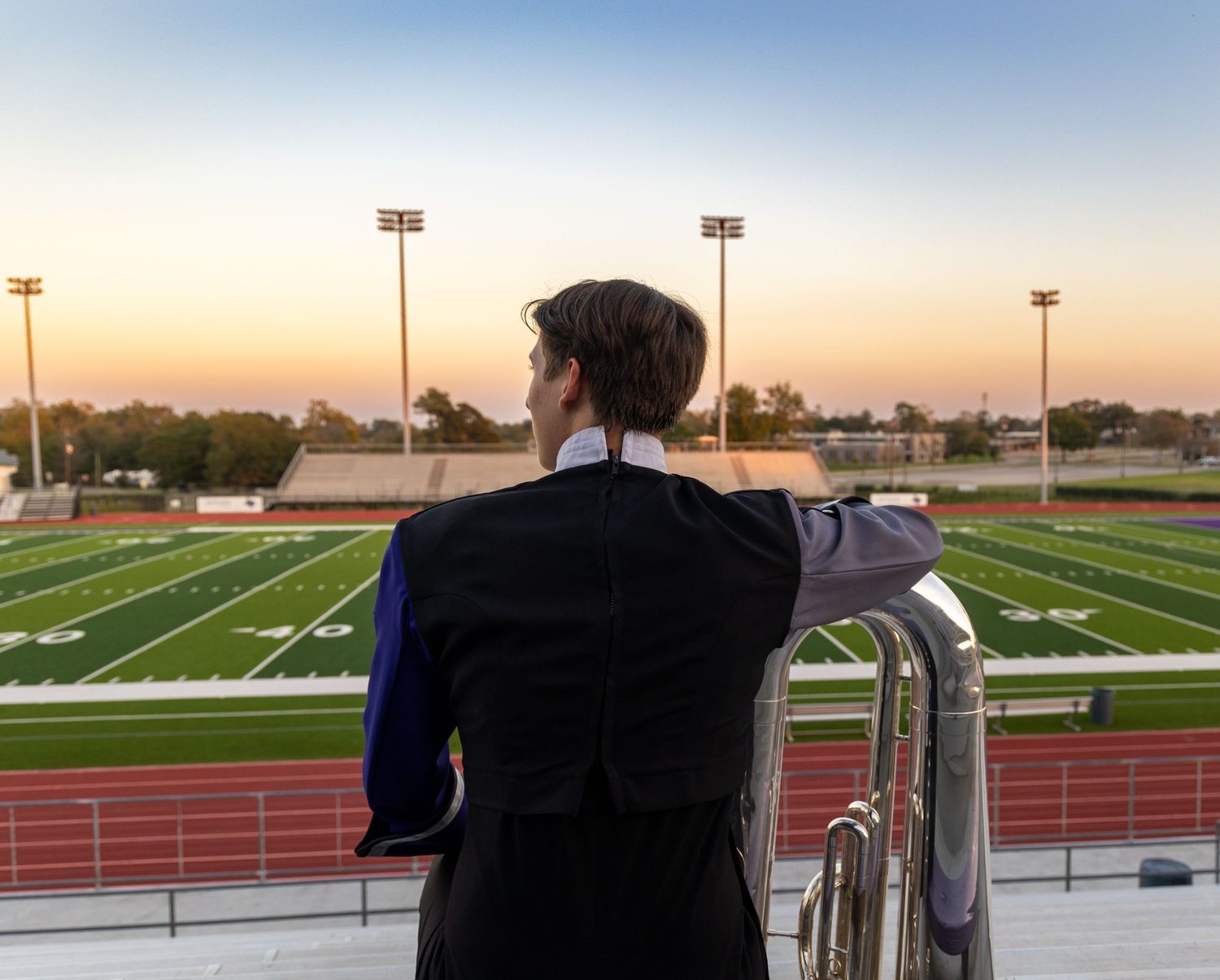 A person in formal attire stands overlooking an empty sports stadium field at sunset with floodlights illuminating the scene.