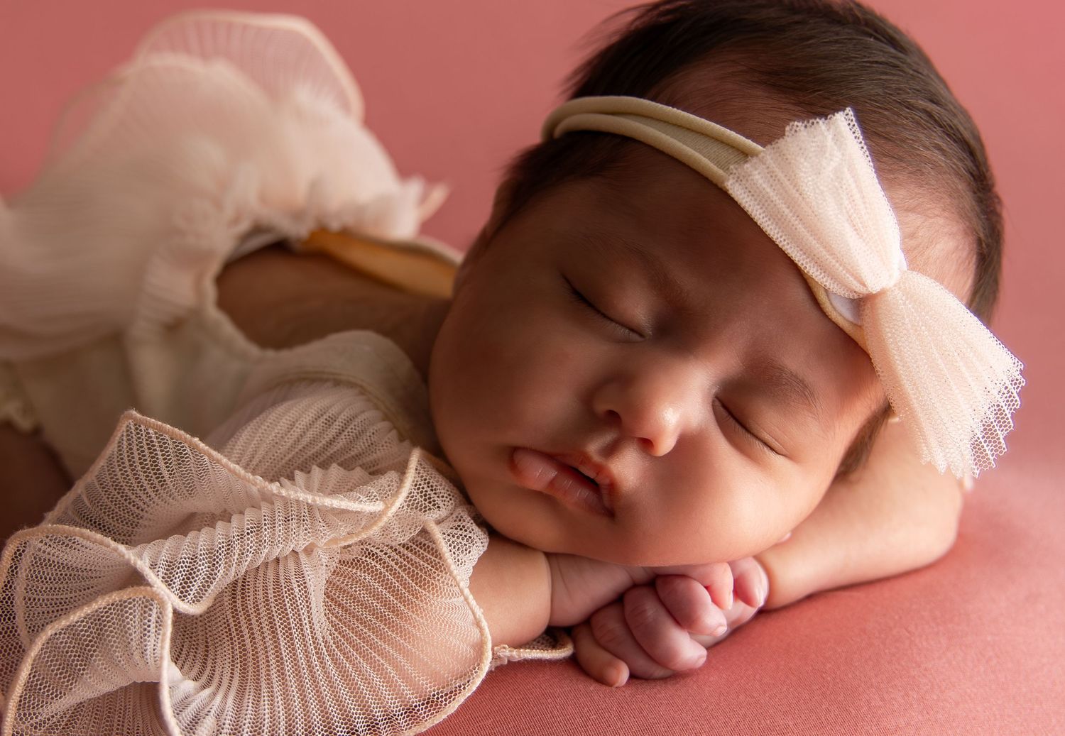 Sleeping newborn baby girl in white lace outfit and bow headband on pink background.