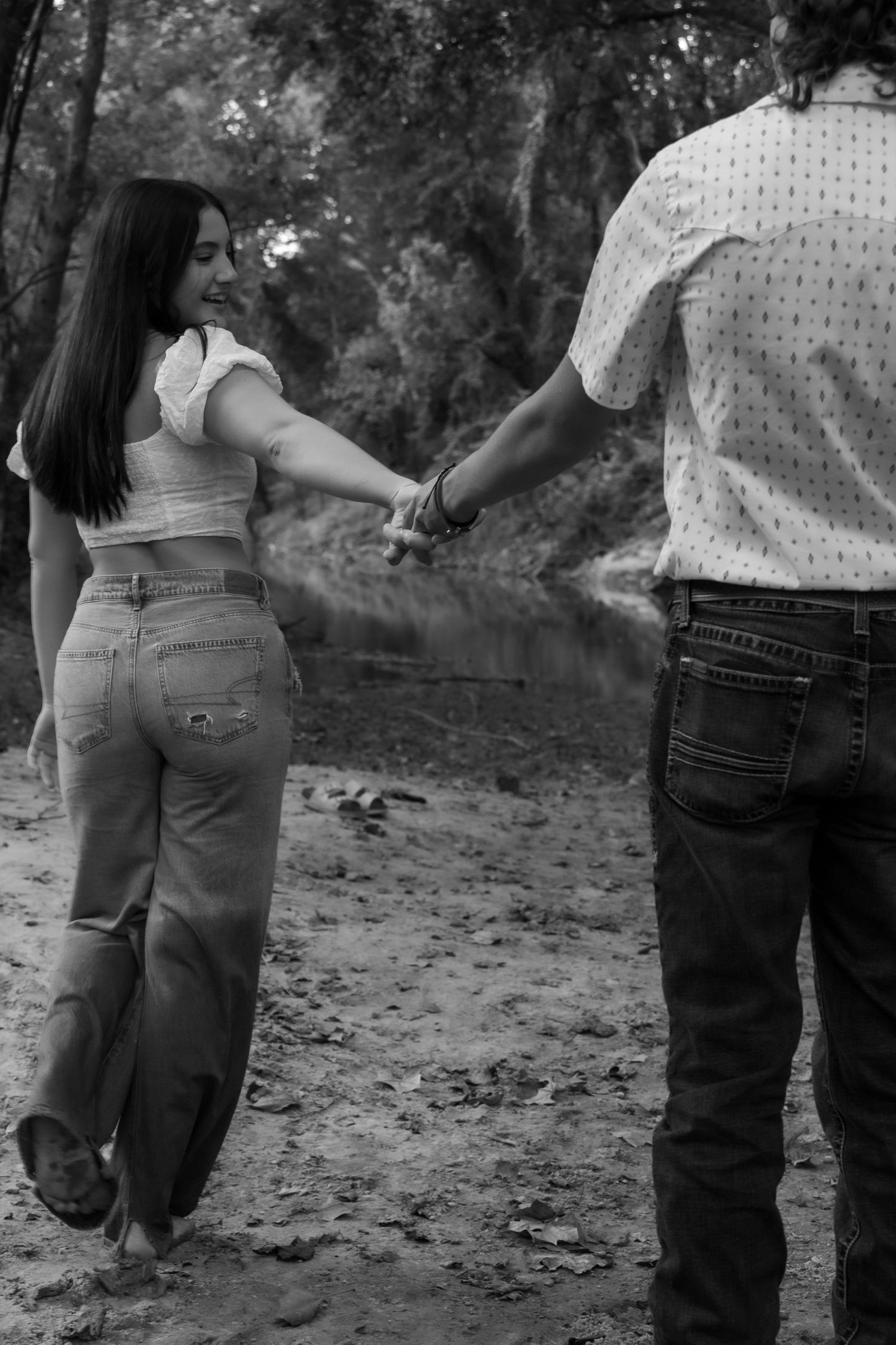 Black and white photo of a couple holding hands while walking on a dirt path through a wooded area.