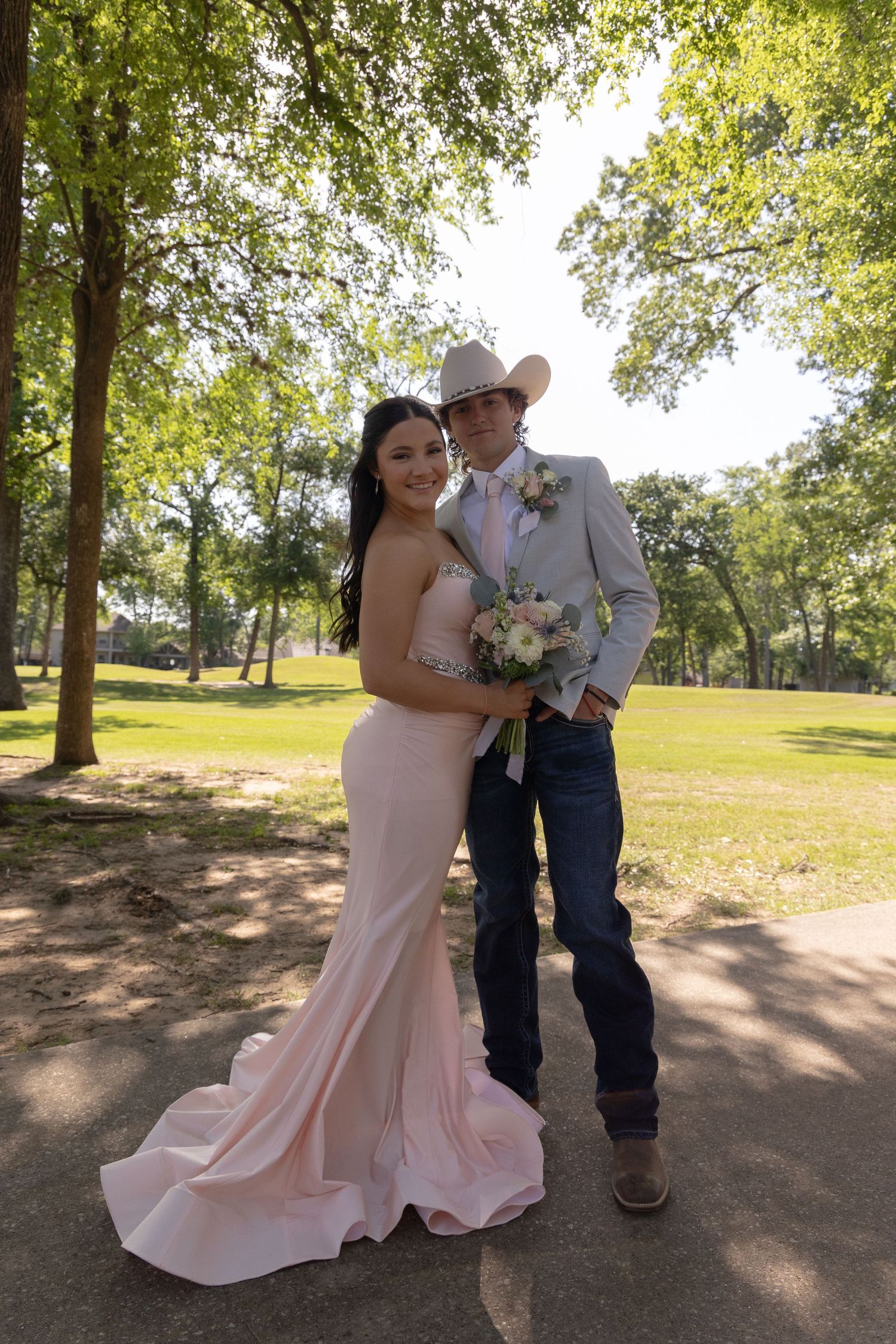 A couple in prom attire poses outdoors, she in a pink gown and he in a gray suit with cowboy hat and boots.