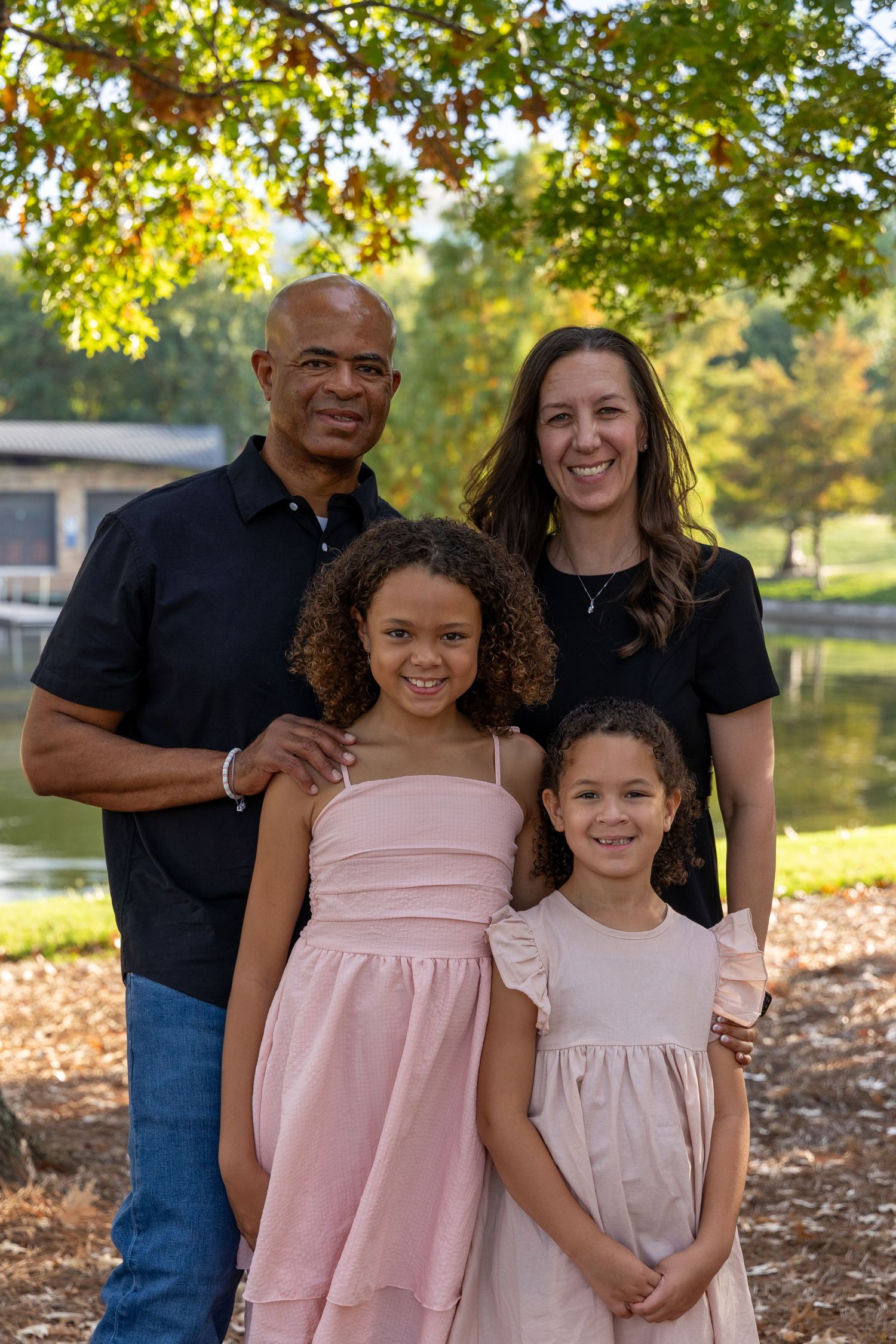 A smiling family of four poses outdoors near a pond, with two girls in pink dresses and their parents in black tops.