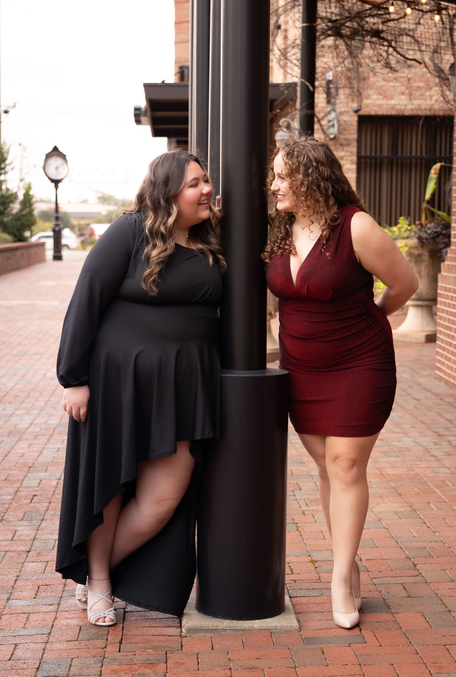 Two plus-size women in elegant dresses pose by a black pole on a brick walkway, smiling at each other.