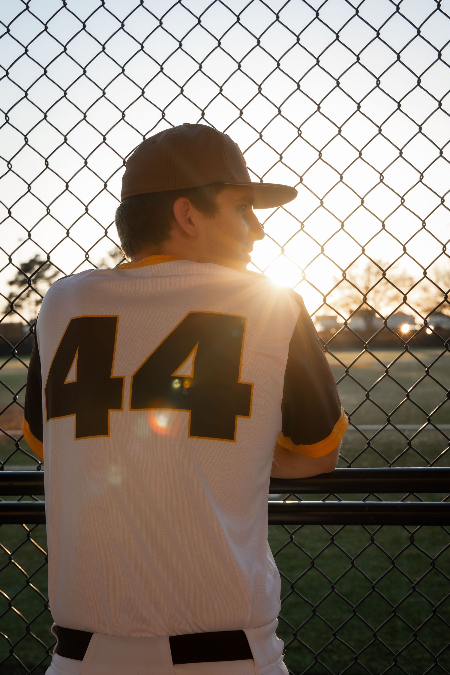 Baseball player wearing jersey number 44 stands at chain-link fence during golden hour sunset on field.