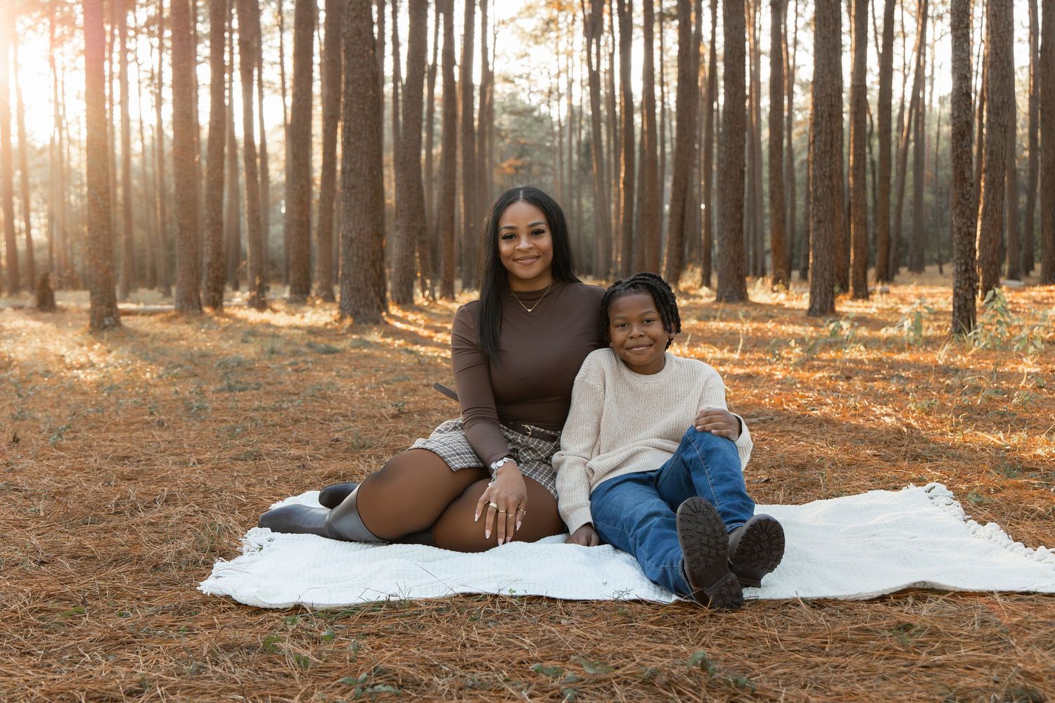 Two women sitting together on a white blanket in a sunlit pine forest during golden hour.