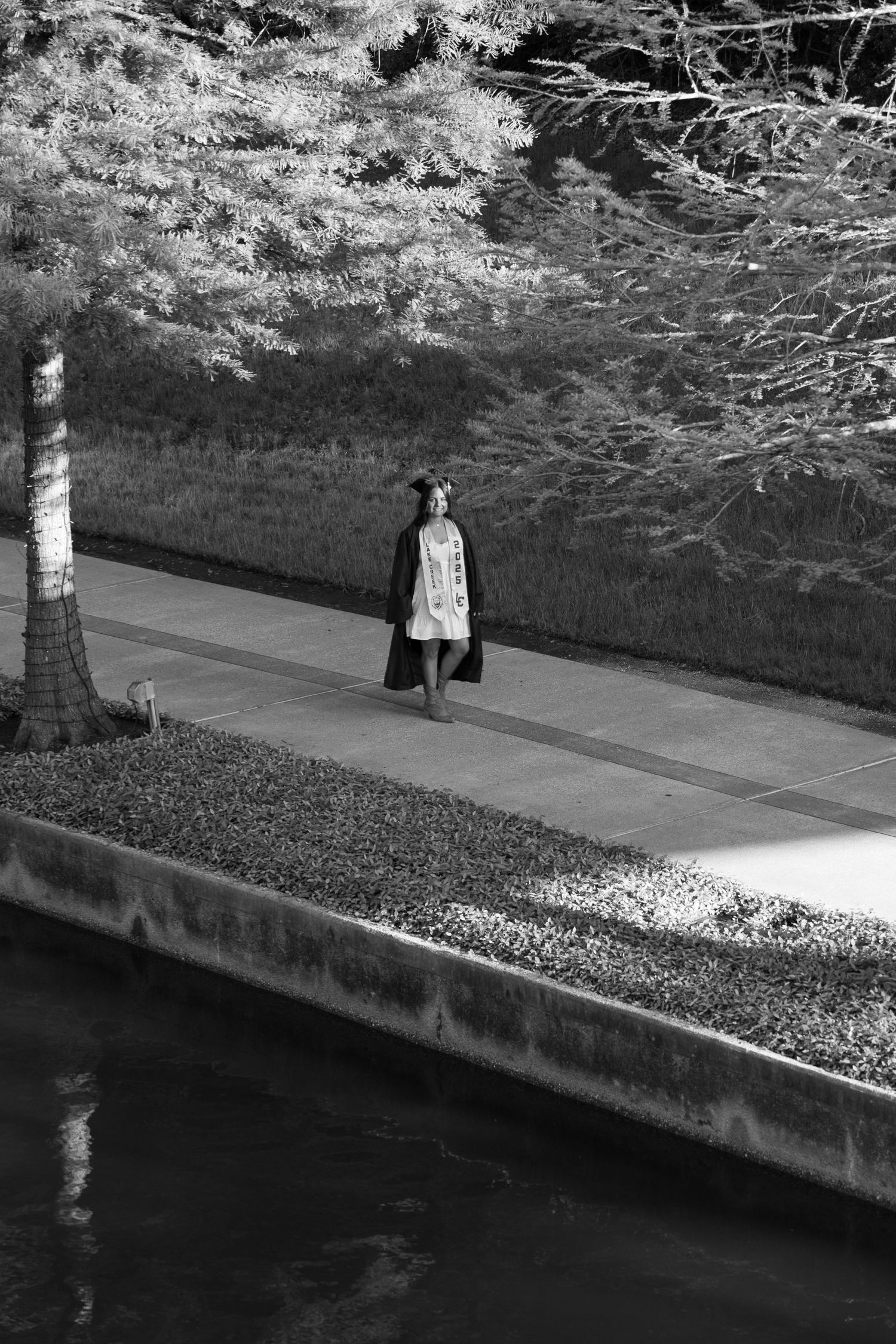 Black and white photo of a woman walking along a serene riverside path lined with trees and manicured hedges.