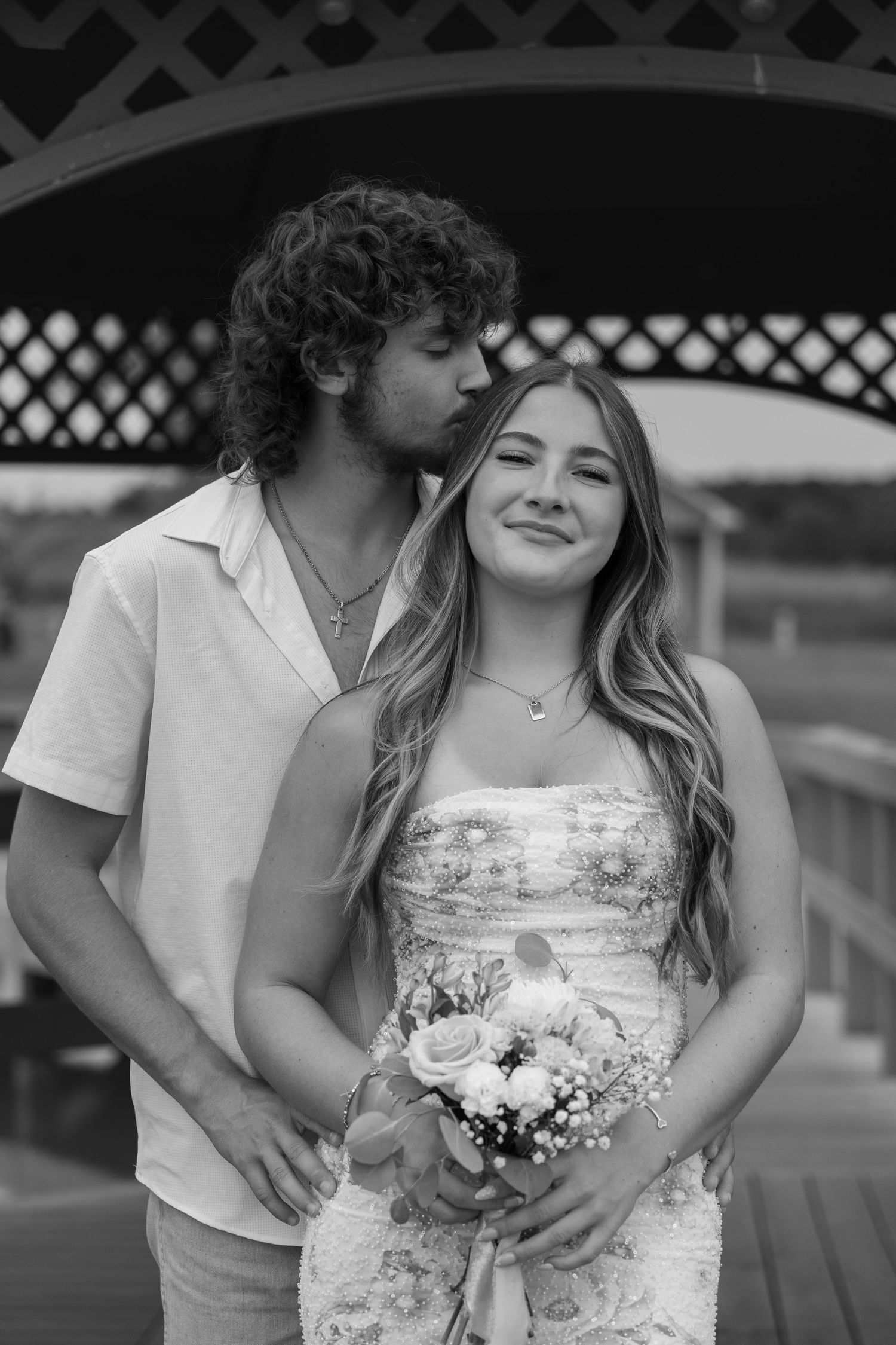 Black and white photo of a couple, man kissing woman's head as she smiles holding a bouquet under a gazebo.