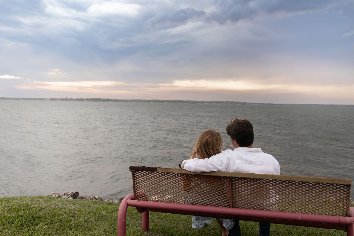 Couple sits closely on a bench overlooking a calm ocean under a cloudy sky at dusk.