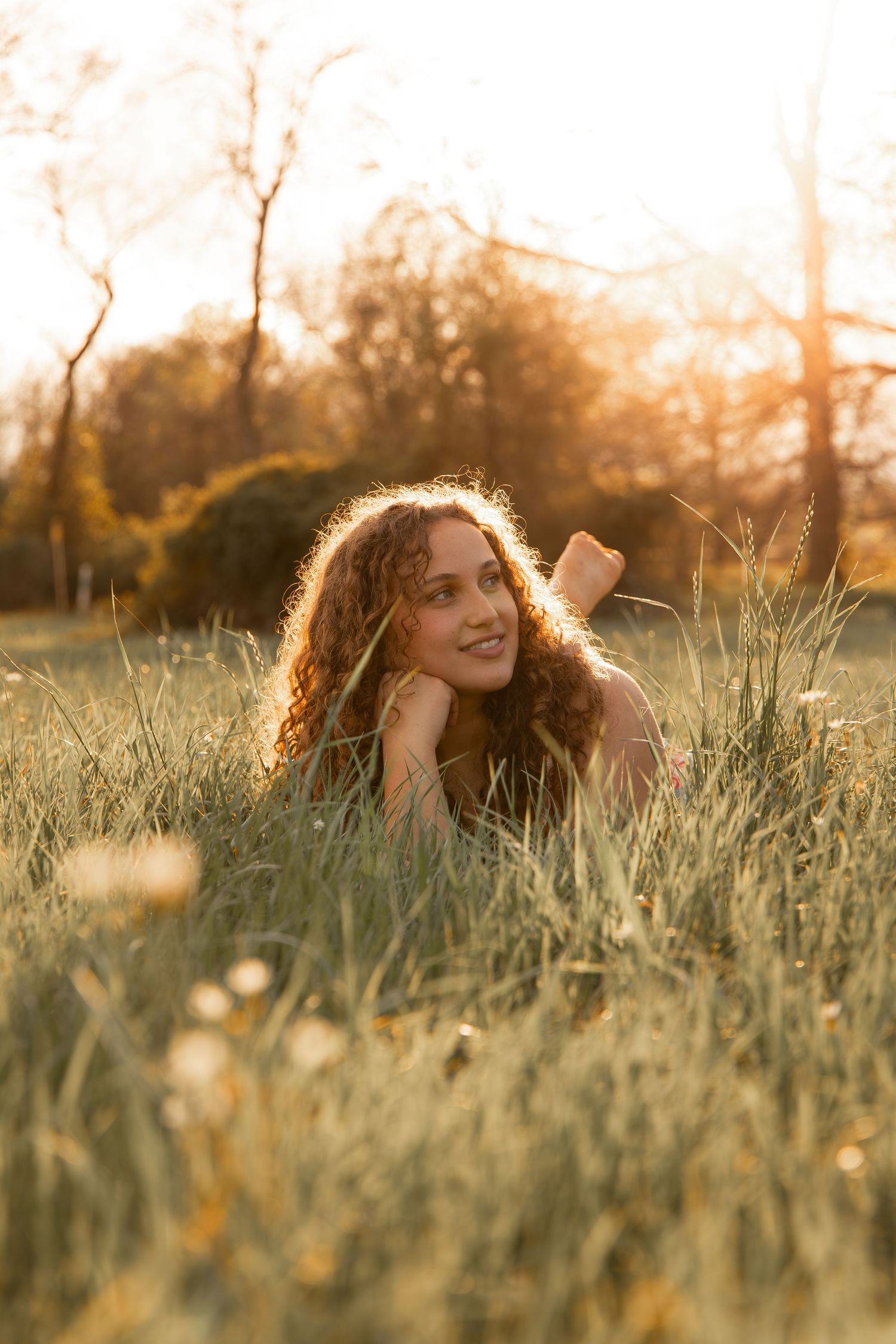 Young woman with curly hair lying in tall grass during golden hour sunset, smiling peacefully in a sunlit meadow.