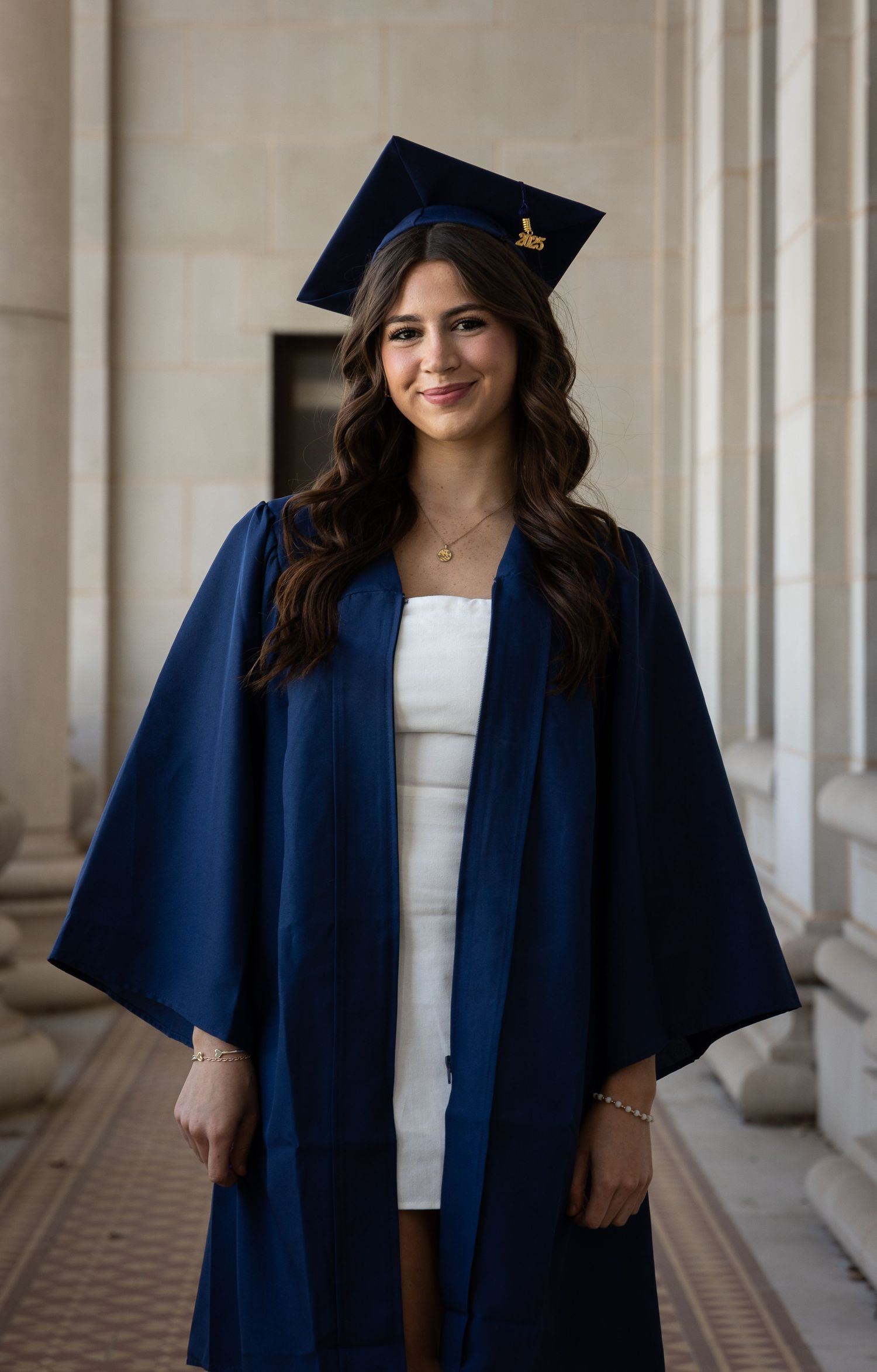 Young woman in navy blue graduation cap and gown smiling between columns, wearing white dress underneath.