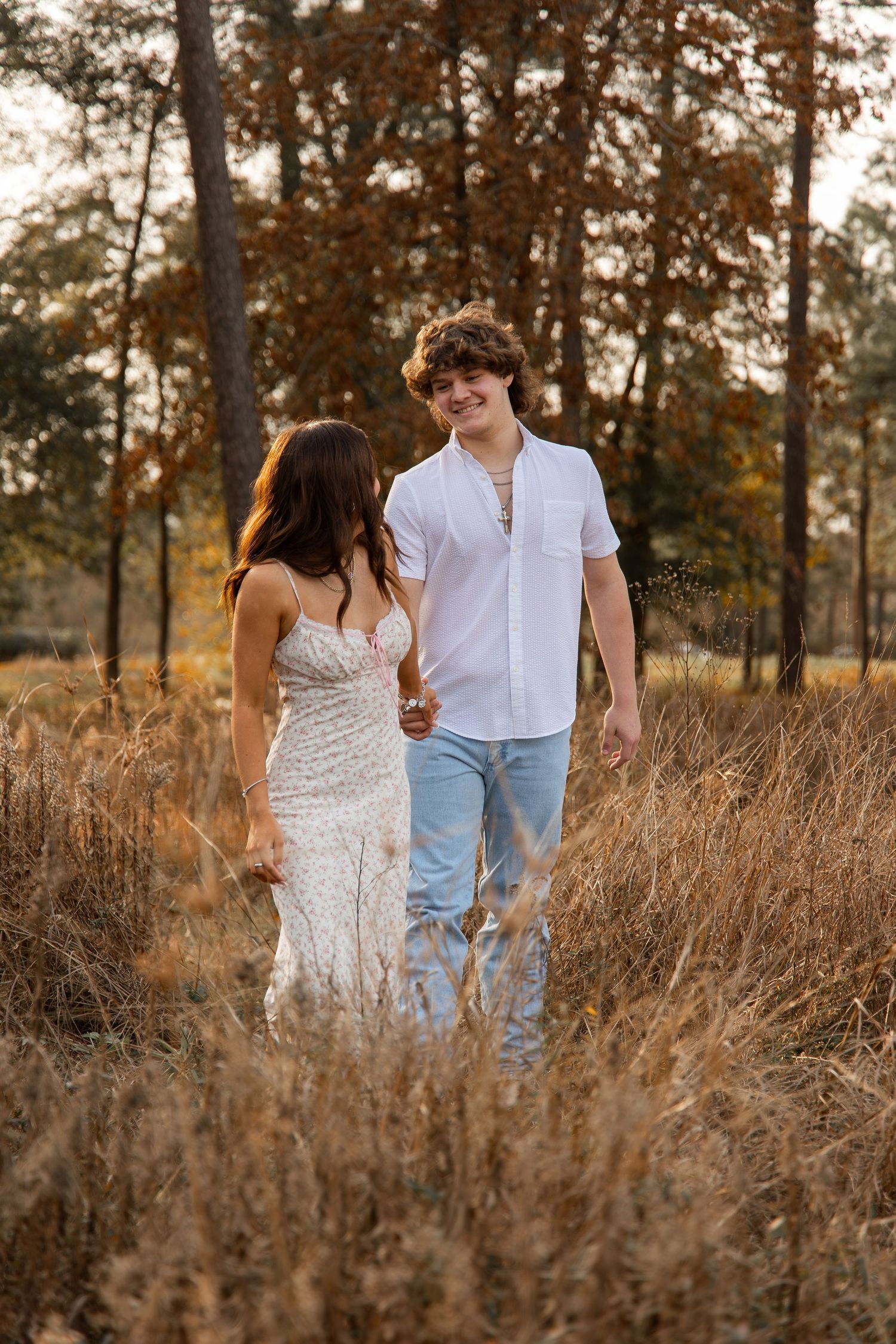 A couple walks hand-in-hand through a golden field, woman in white lace gown, man in white shirt and jeans, autumn trees behind.