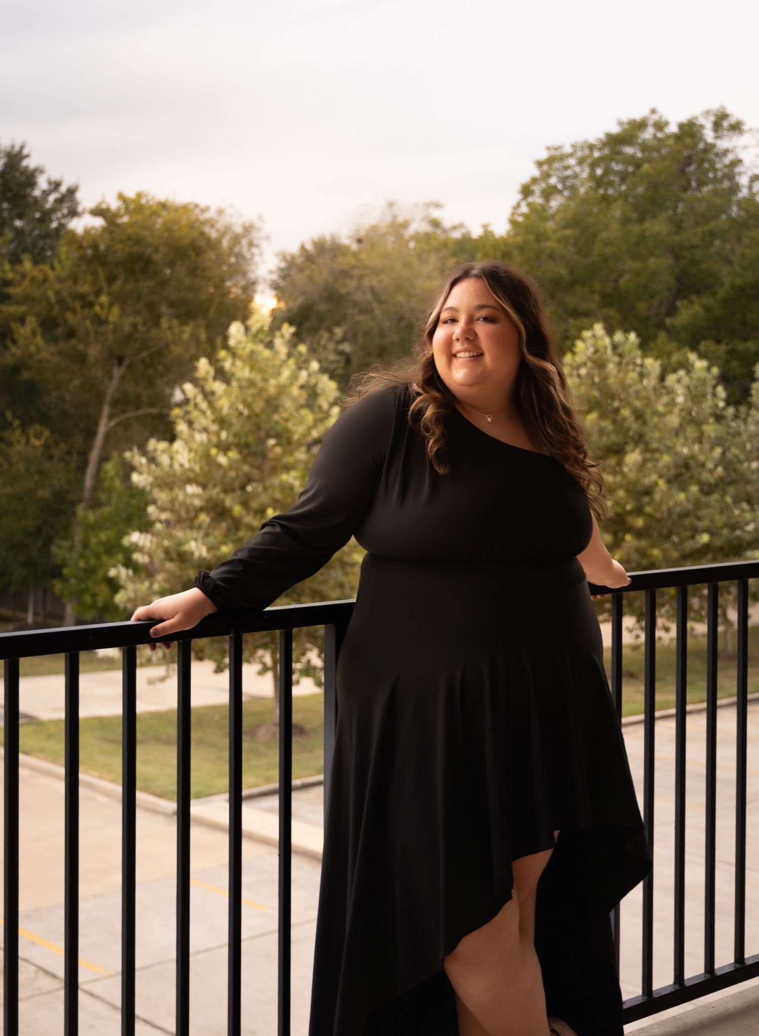 Woman in black asymmetrical dress posing on balcony with lush green trees in background at golden hour.