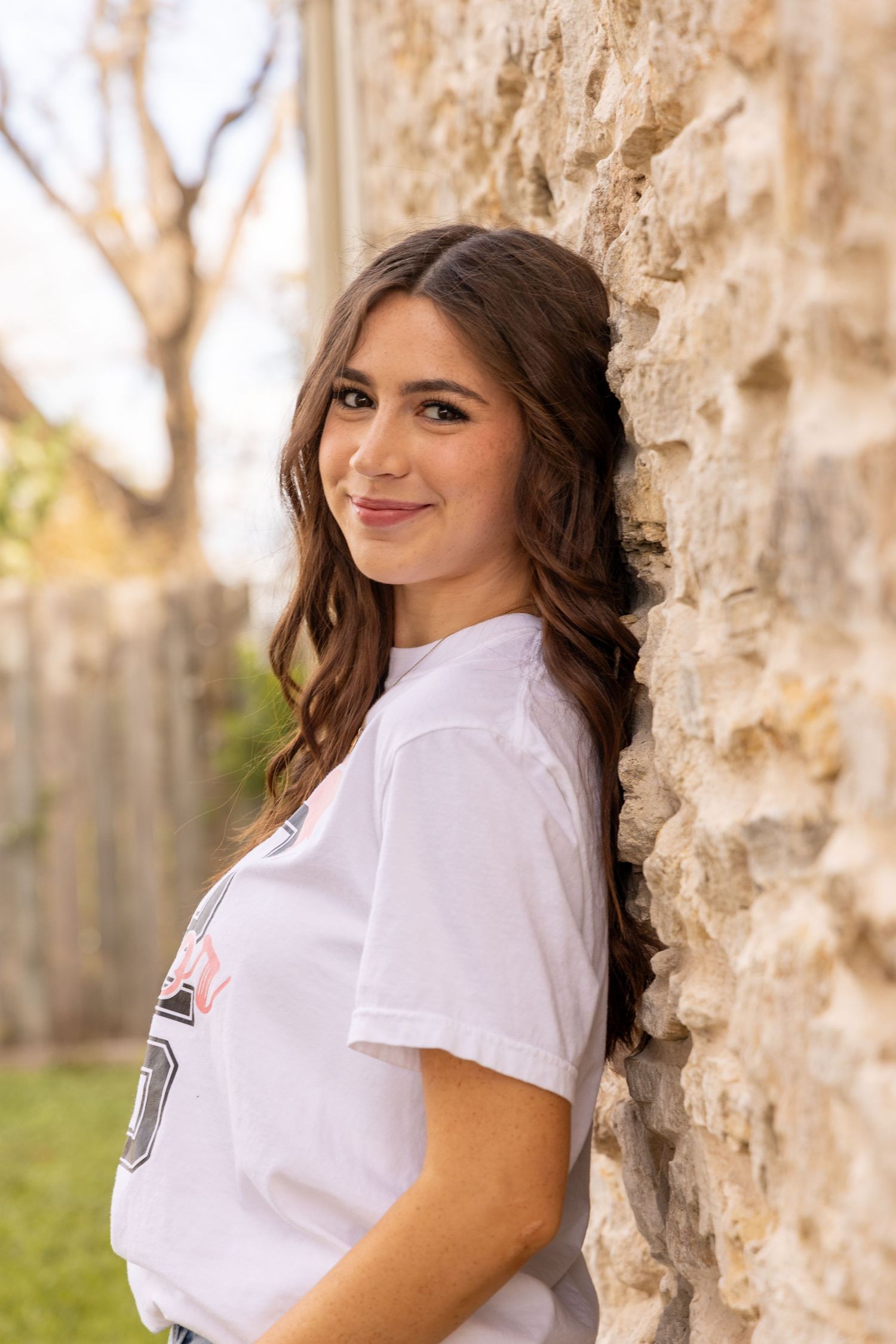 A young woman in a white t-shirt leans against a textured stone wall while standing outdoors in natural lighting.