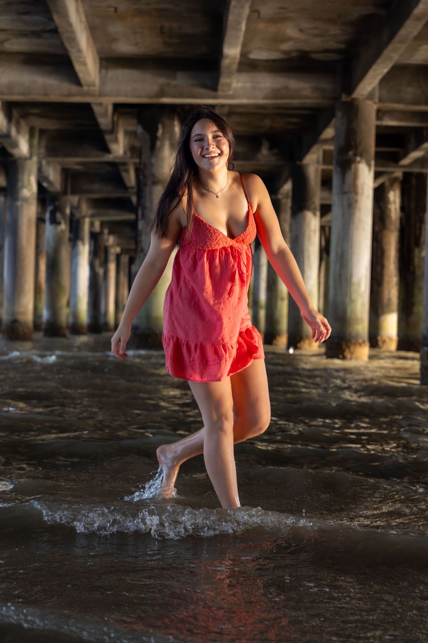 Young woman in red dress walking barefoot through ocean waves under a wooden pier, smiling joyfully.