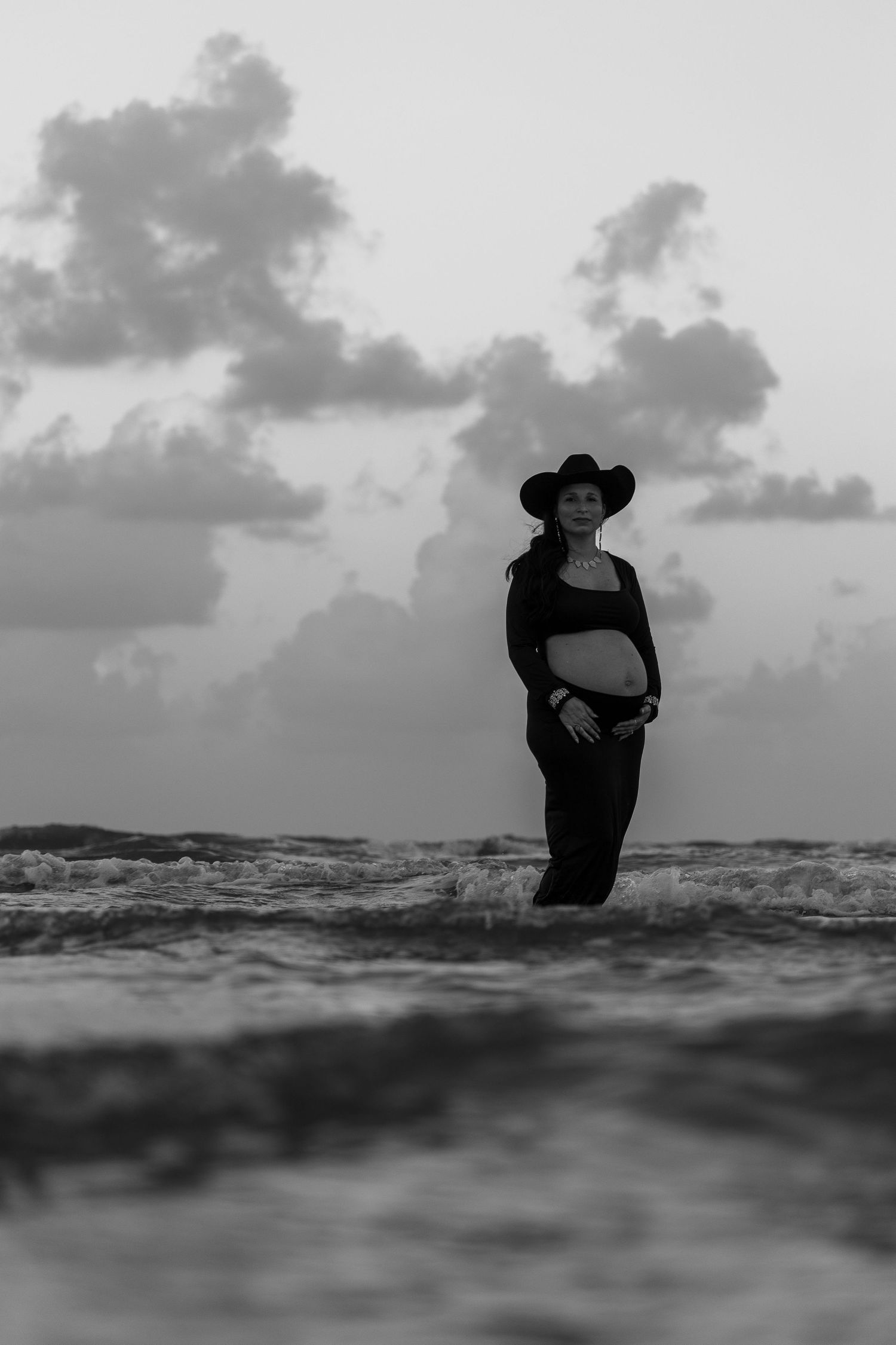 Black and white photo of pregnant woman in cowboy hat standing in ocean waves at sunset.