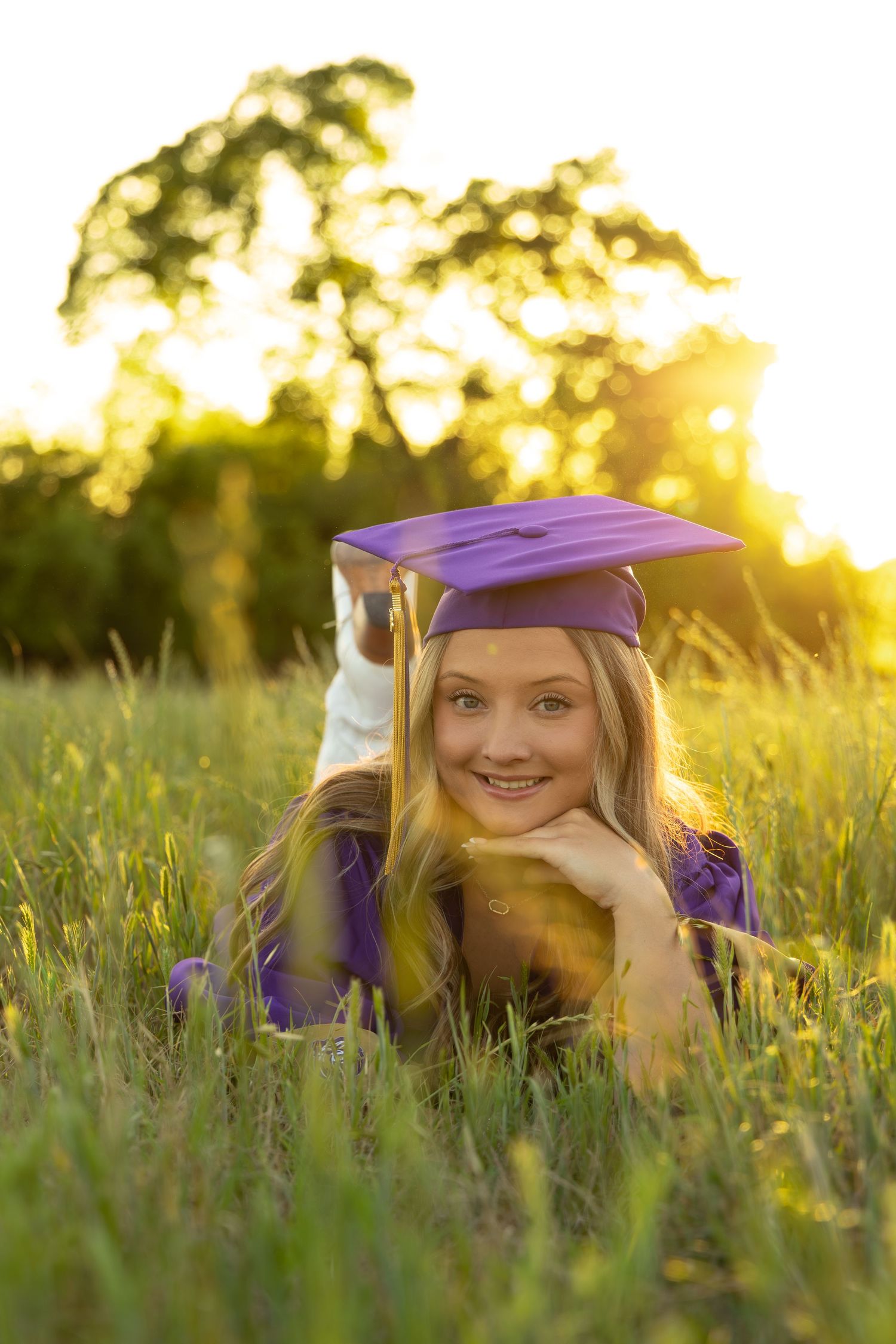 Blonde graduate in purple cap and gown lying in golden sunlit grass field smiling at camera.