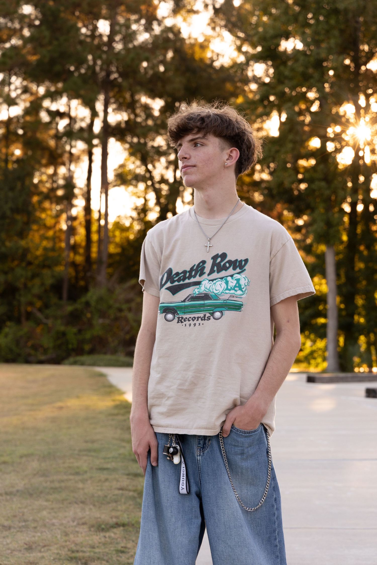 Young man in Death Row Records tee and jeans poses outdoors at golden hour with trees in background.