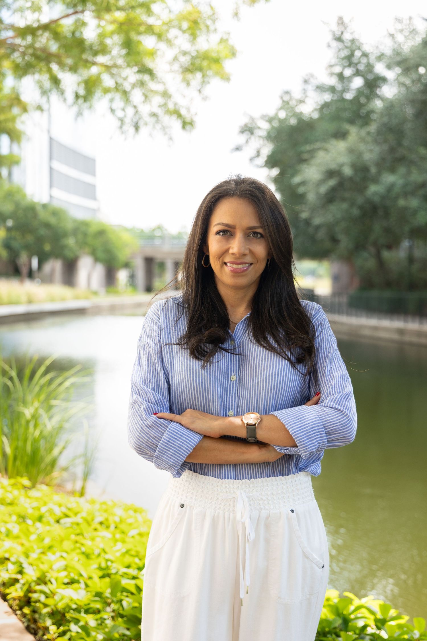 Professional woman in blue striped shirt and white pants standing outdoors near a pond with arms crossed.