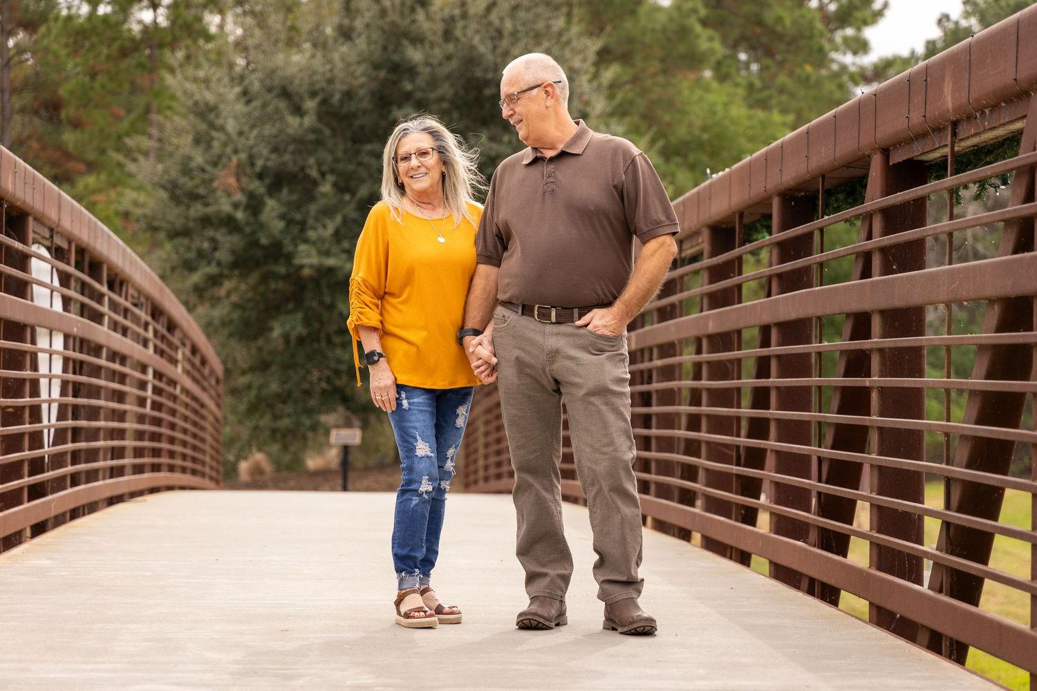 Couple holding hands and smiling while walking on a wooden bridge surrounded by lush green trees.