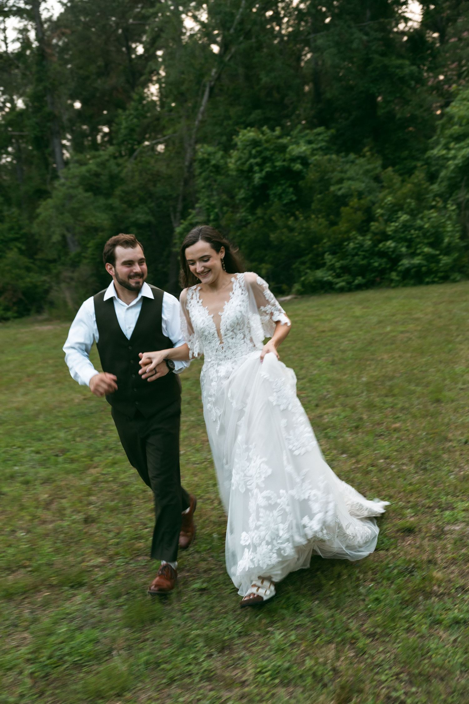 A couple runs joyfully across a grassy lawn, with the bride wearing a lace wedding gown and groom in a vest and slacks.