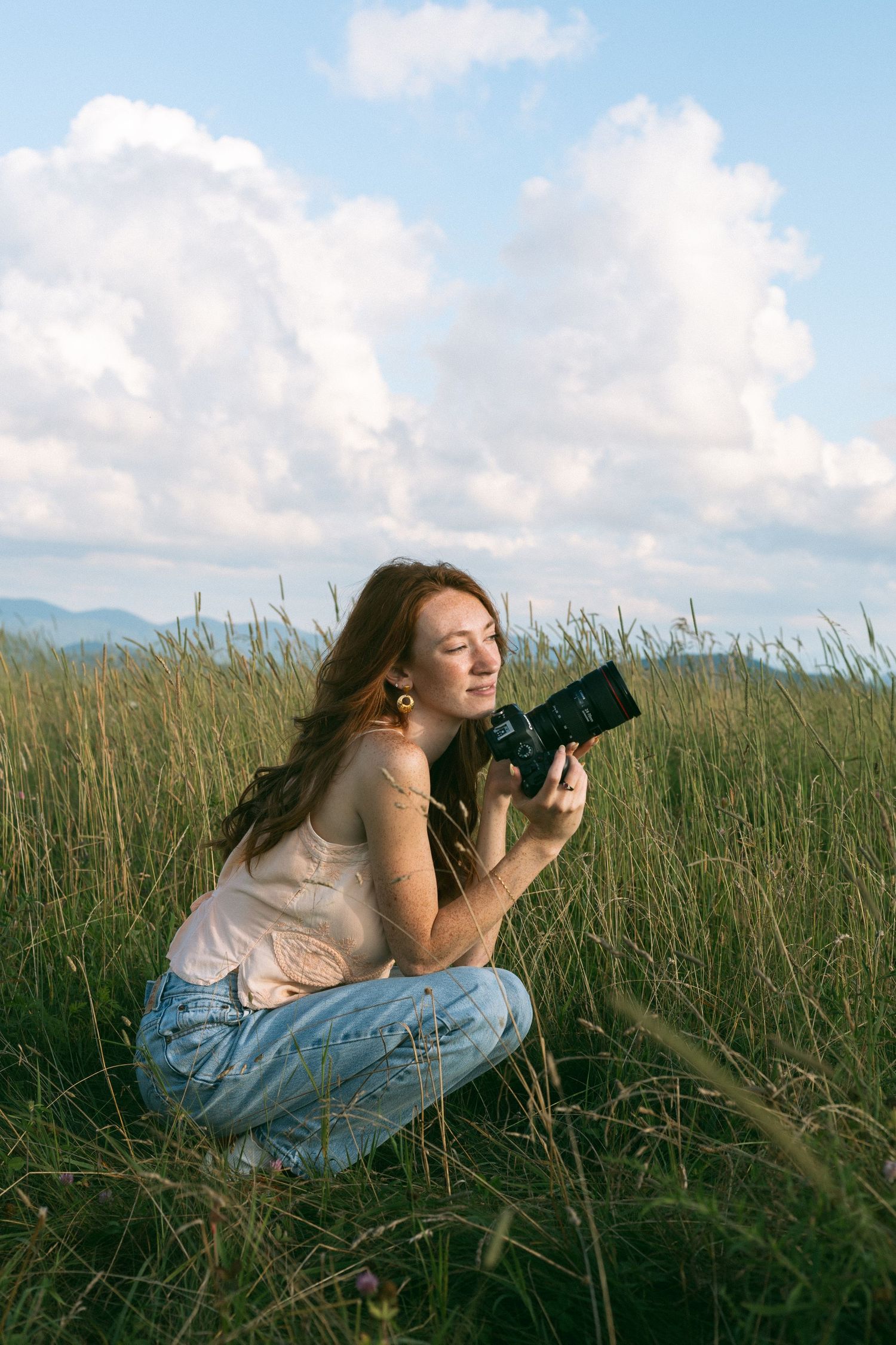 A photographer in a white top lies in tall grass taking photos with a professional camera on a sunny day with blue skies.